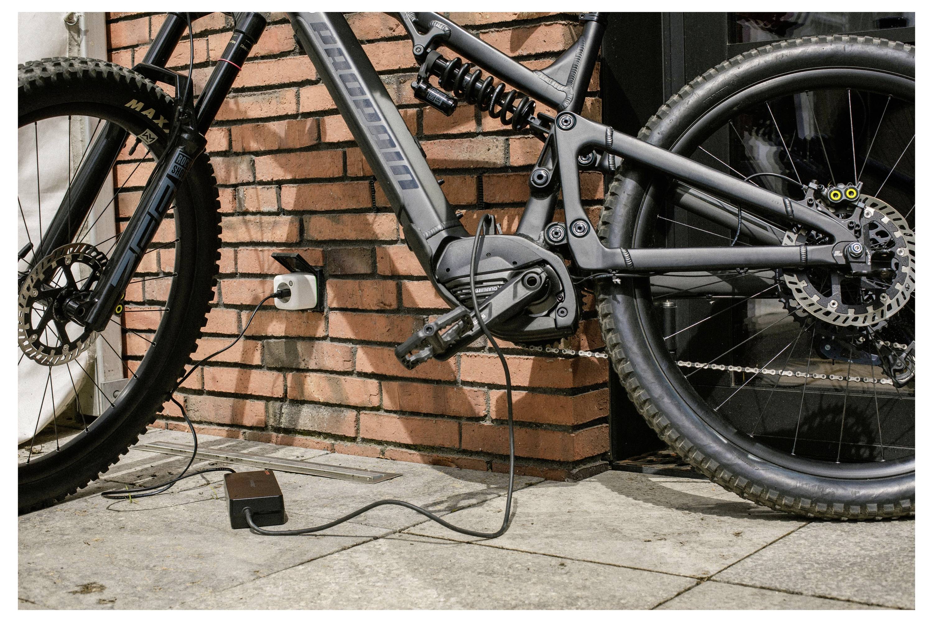 Elektrisches Fahrrad, das an eine Steckdose angeschlossen ist und auf einer Terrasse mit Backsteinmauer im Hintergrund aufgeladen wird.