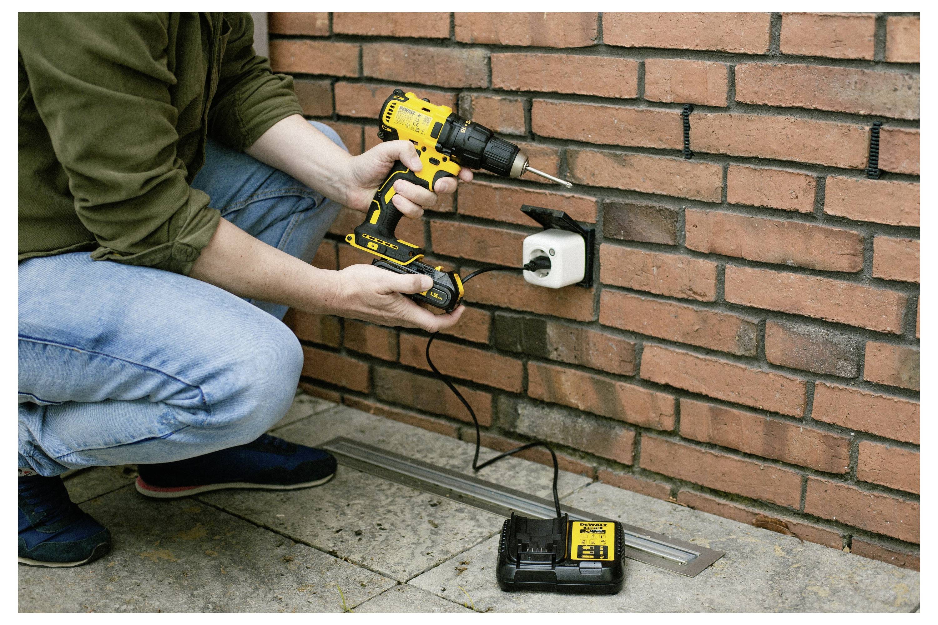 Eine Person benutzt einen gelben kabellosen Bohrer, um ein Gerät an einer Ziegelmauer zu befestigen, was DIY-Heimverbesserungen hervorhebt. Eine Batterie und ein Ladegerät sind auf dem Boden sichtbar.