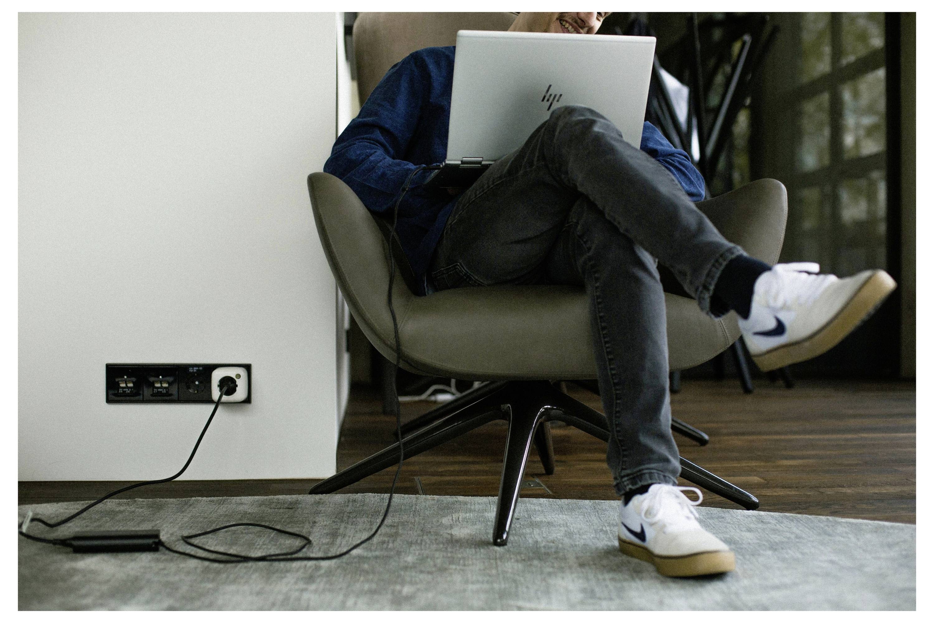 Person, der in einem Stuhl sitzt und einen Laptop benutzt, mit überkreuzten Beinen. Eine Steckdosenleiste liegt auf dem Boden und ist in eine Wandsteckdose gesteckt. Lässige Inneneinrichtung.