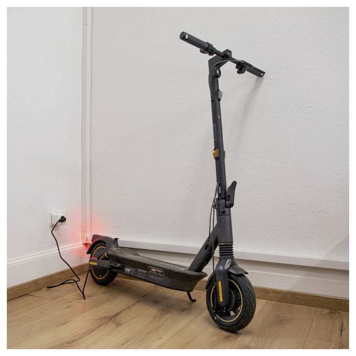 Ein Elektroroller steht drinnen zum Laden, mit einer sichtbaren roten Leuchtanzeige. Er lehnt an einer weißen Wand auf einem Holzboden.