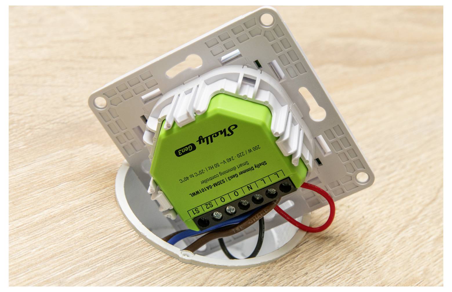 Ein grünes intelligentes Schaltermodul mit verbundener Verkabelung, montiert auf einer rechteckigen Wandplatte, platziert auf einer Holzoberfläche.