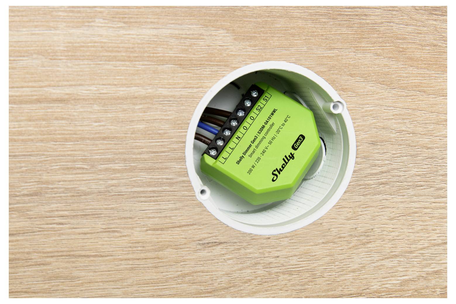 Ein grünes Shelly 1PM Smart-Relais ist in einer Elektrobox installiert, sichtbar vor einer Holzoberfläche, das seine Verdrahtungsverbindungen zeigt.