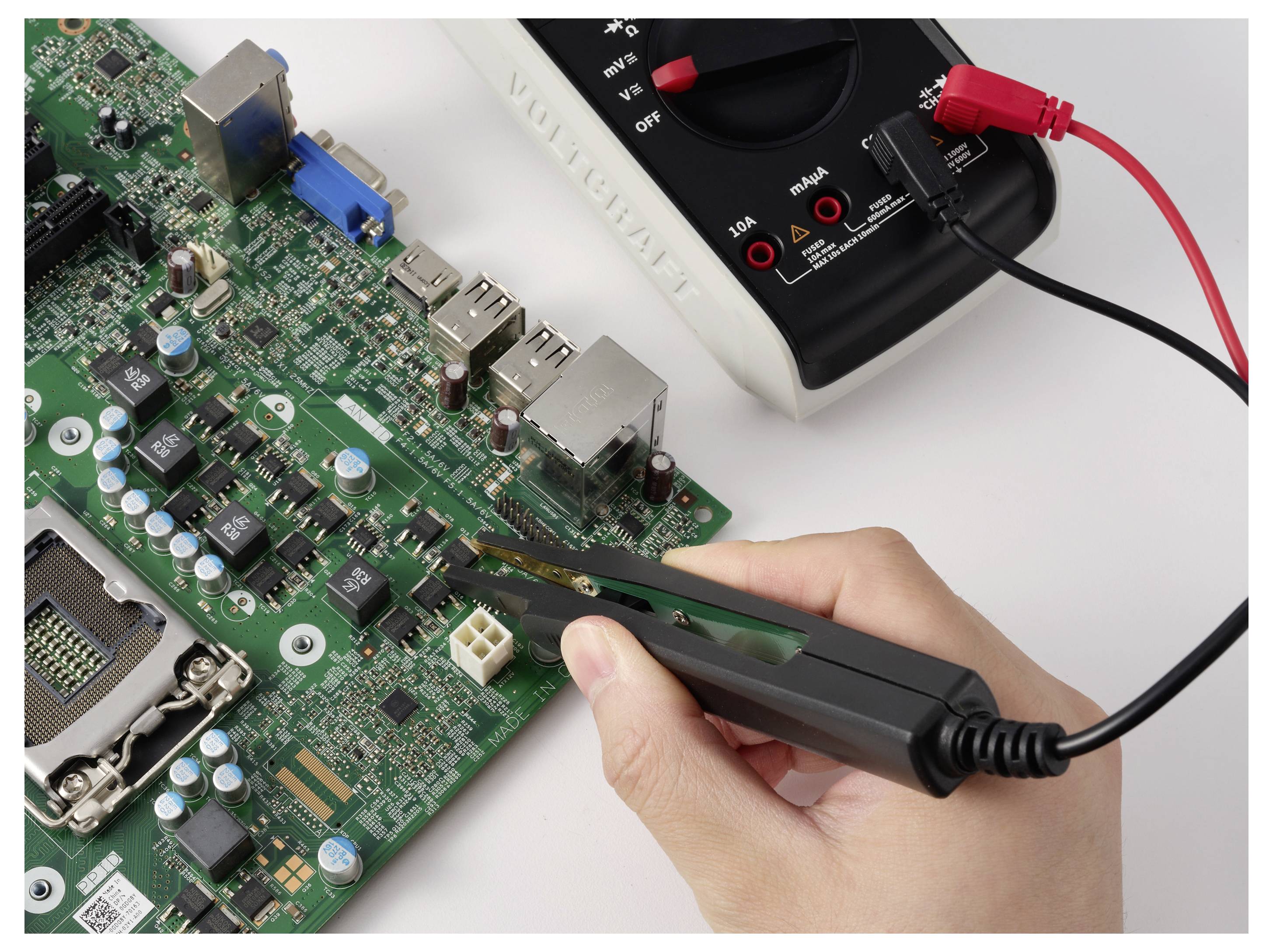 Hand hält eine Pinzette und untersucht eine Leiterplatte, während ein Multimeter mit roten und schwarzen Messleitungen die Komponenten prüft.