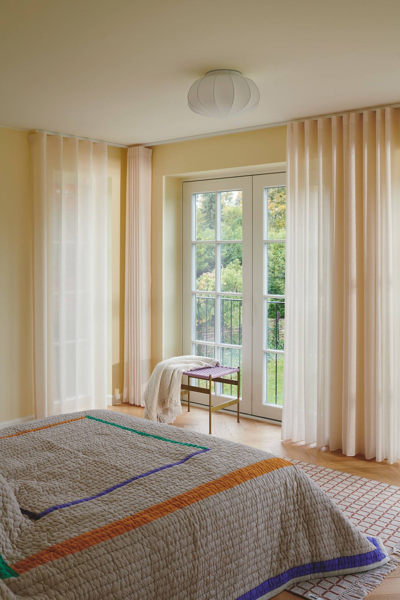 Ein gemütliches Schlafzimmer mit einem großen Fenster, leichten Vorhängen und einer farbenfrohen gesteppten Bettdecke. Ein kleiner Tisch hält eine gefaltete Decke.