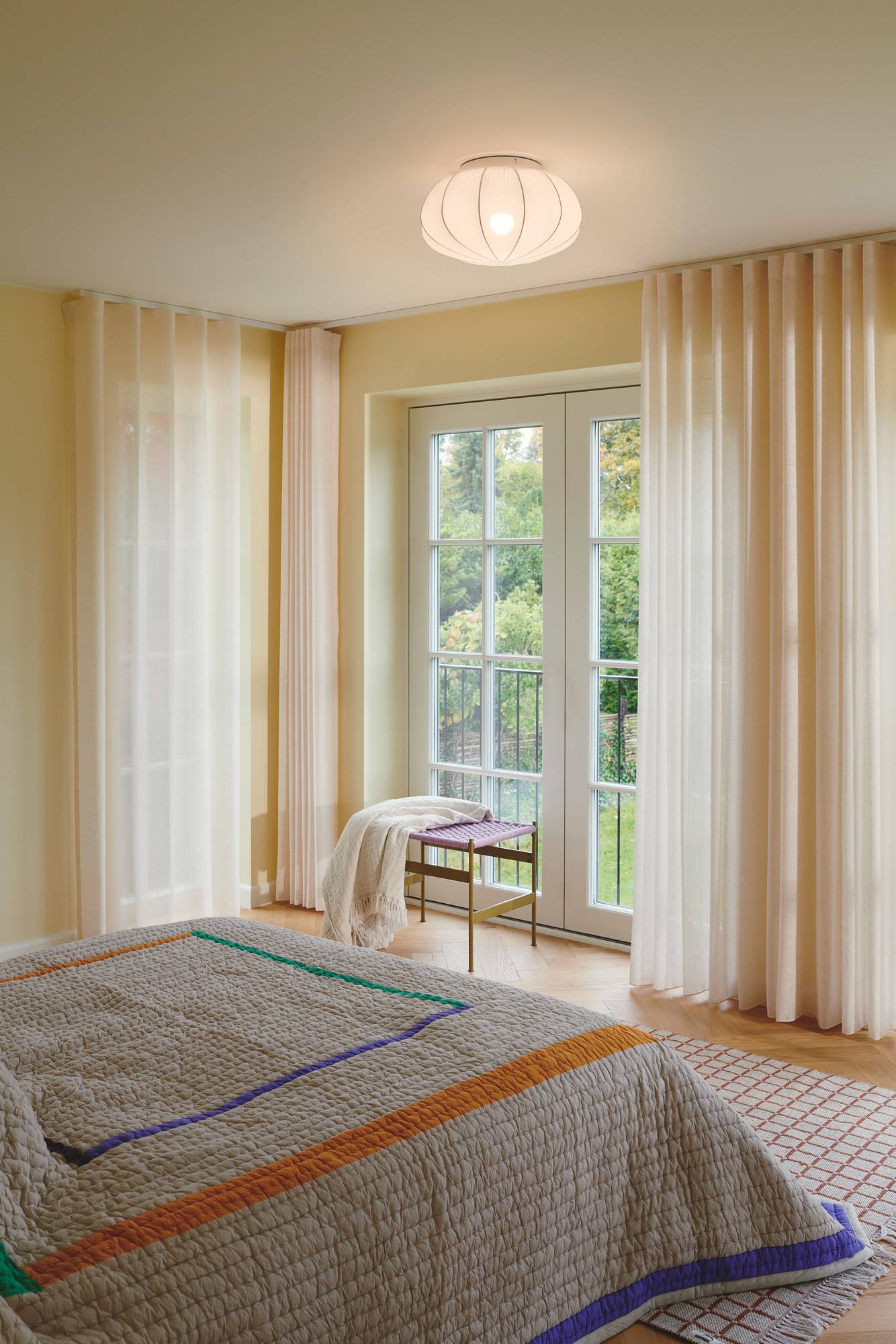 Ein gemütliches Schlafzimmer mit einem gesteppten Bett, transparenten Vorhängen und Glastüren, die den Blick auf einen Garten ermöglichen und eine ruhige Atmosphäre schaffen.