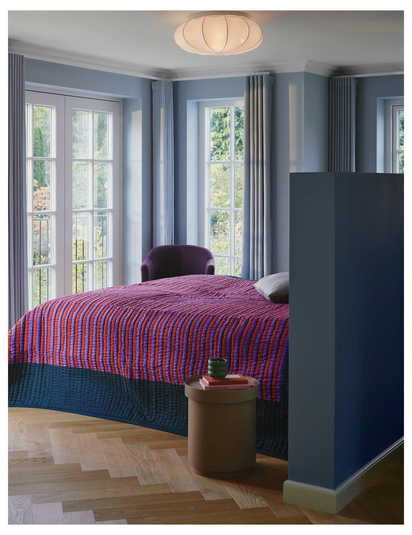 Ein gemütliches Schlafzimmer mit großen Fenstern verfügt über ein Bett, das mit einer farbenfrohen gestreiften Steppdecke bedeckt ist. Ein violetter Stuhl steht neben den Fenstern.