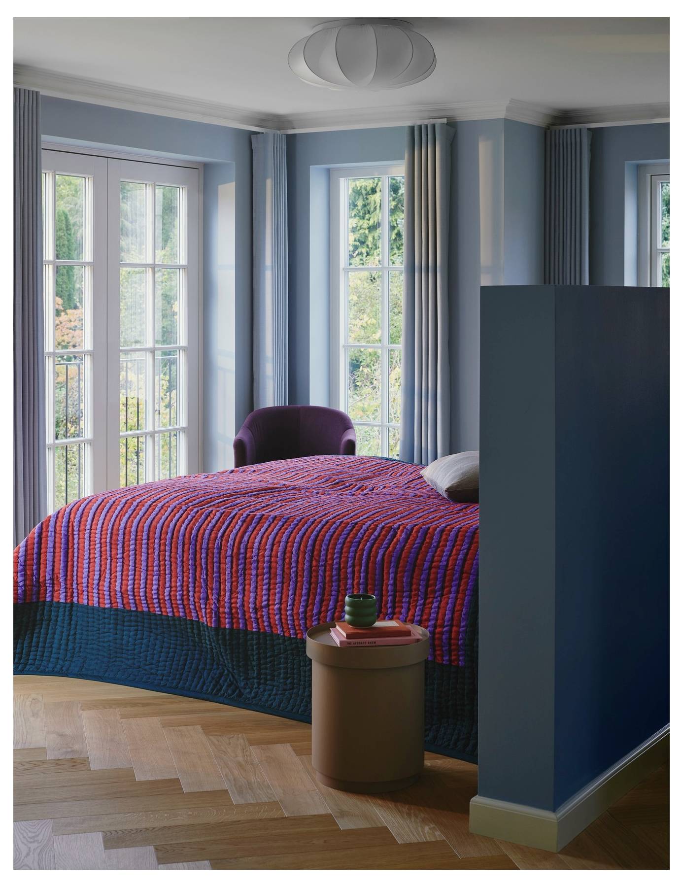 Ein gemütliches Schlafzimmer mit einem lila Sessel, einem Bett mit einer lebendigen rot-blauen gemusterten Bettdecke und großen Fenstern mit Blick auf Grünflächen.