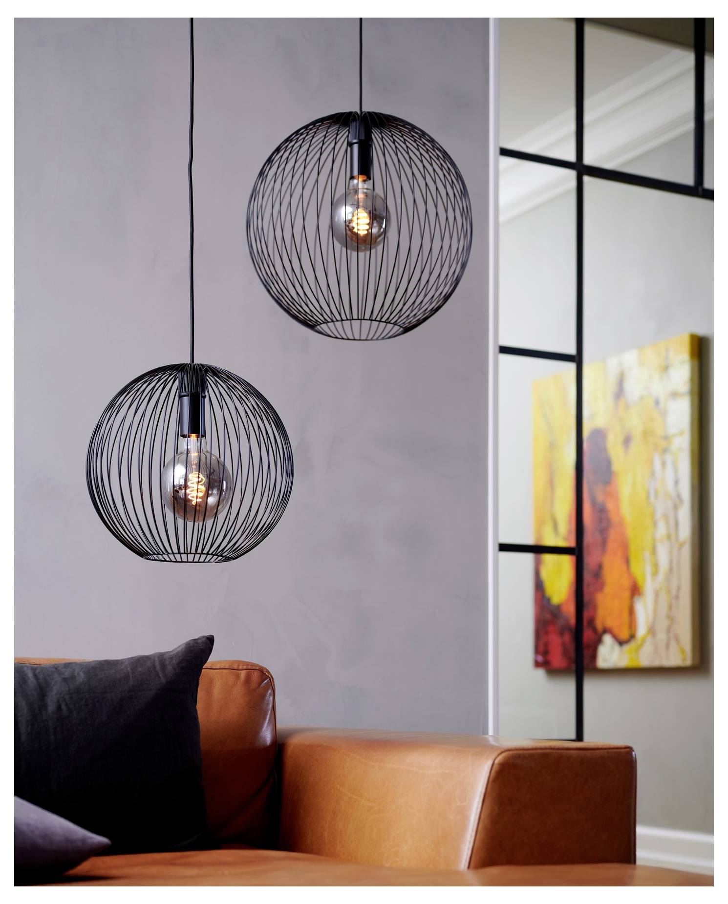 Zwei kugelförmige Draht-Pendelleuchten hängen über einem braunen Ledersofa mit schwarzen und orangefarbenen Kissen. Ein farbiges abstraktes Gemälde befindet sich im Hintergrund.