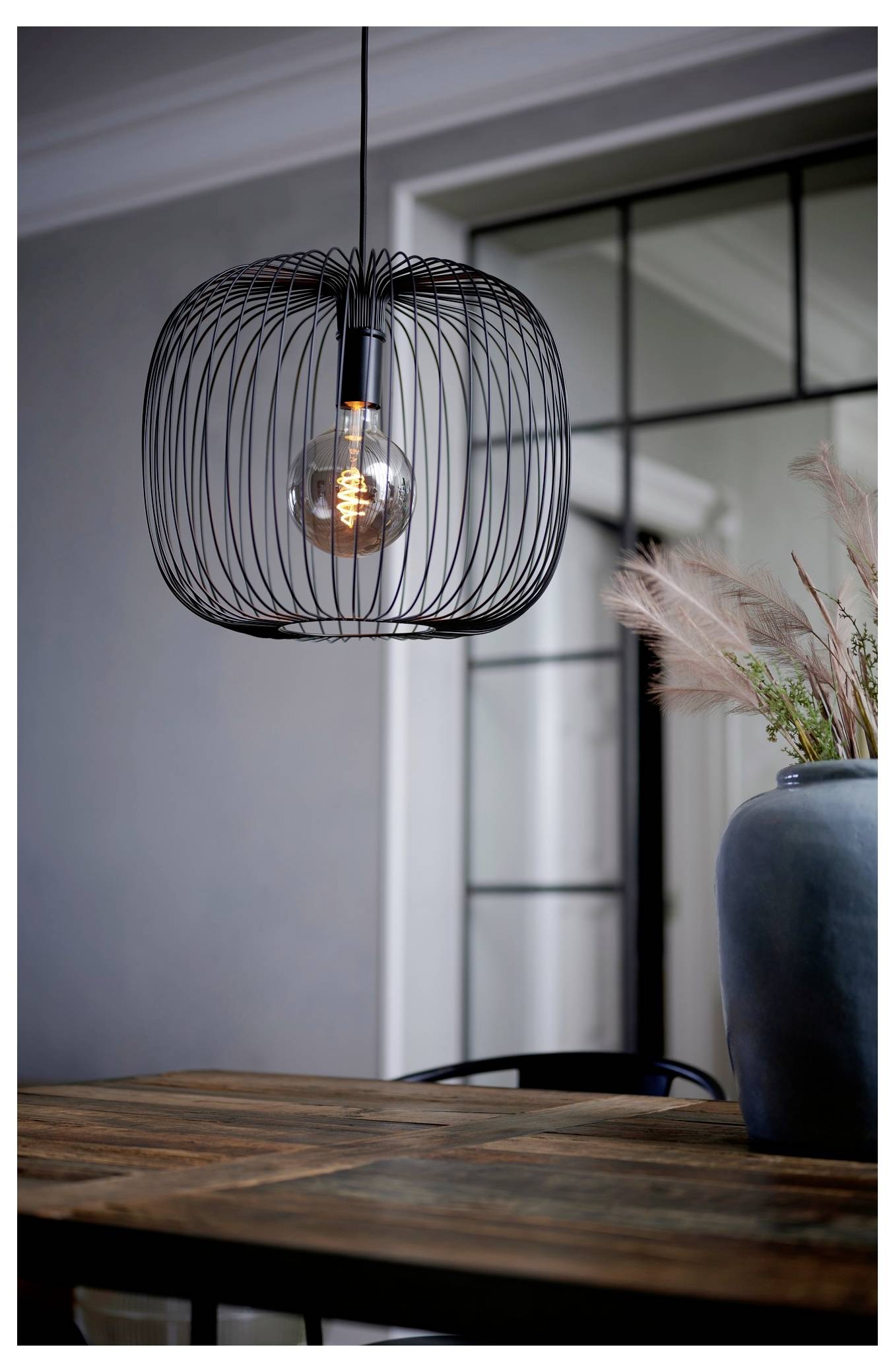 Eine moderne, kugelförmige Drahtpendelleuchte hängt über einem Holztisch. Eine blaue Vase mit getrockneten Pflanzen steht auf dem Tisch, neben einem Fenster.