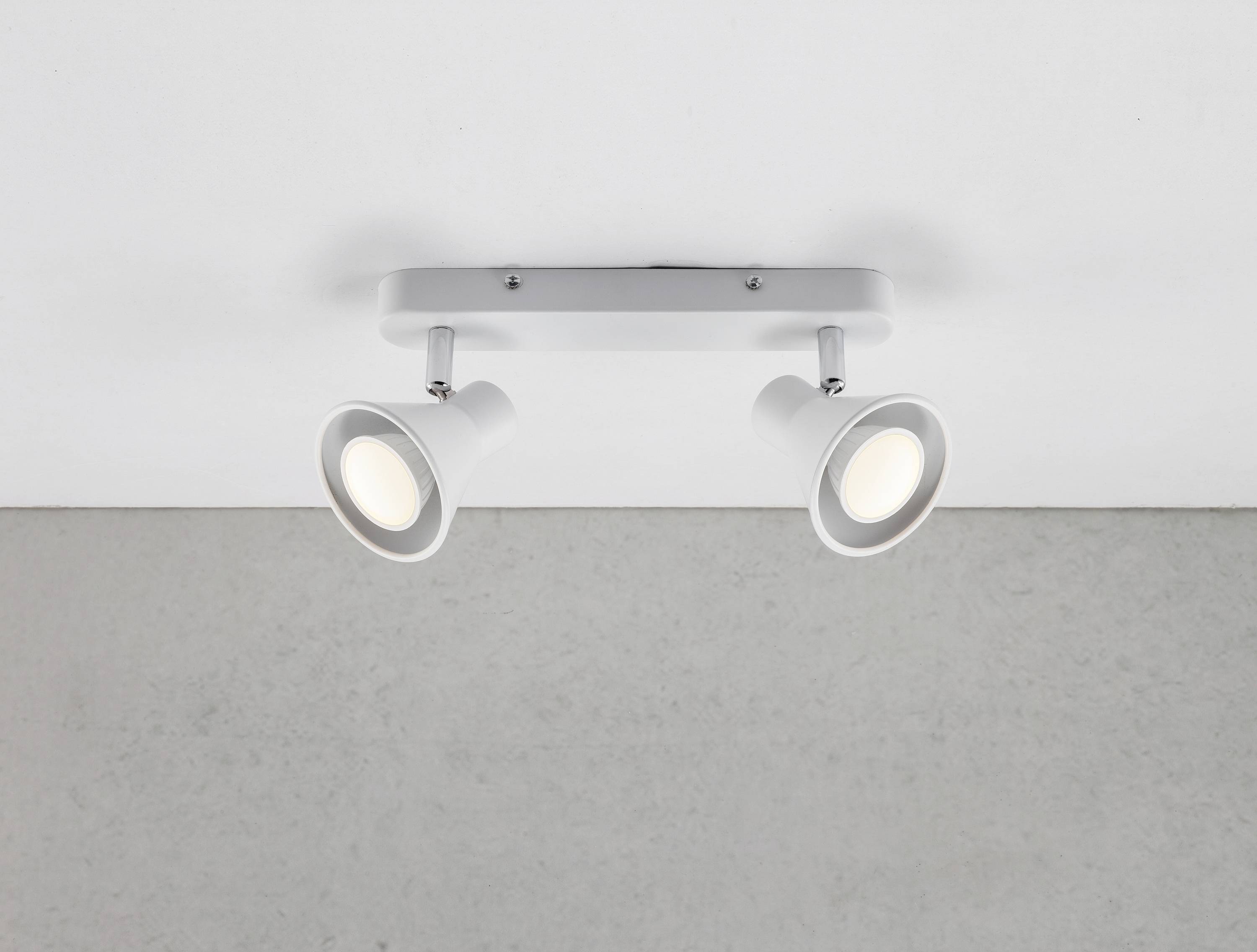 An der Decke montierte, duale Spotlightleuchte mit weißen Lampen, die eine grau strukturierte Wand beleuchtet.