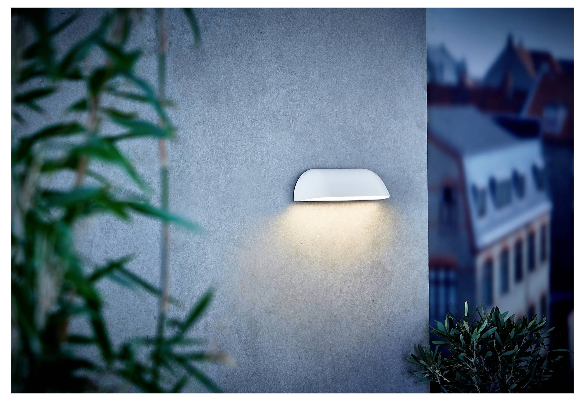 Eine moderne weiße Wandleuchte beleuchtet eine strukturierte graue Wand, mit unscharfem Grün und Gebäuden im Hintergrund, was eine ruhige Atmosphäre schafft.