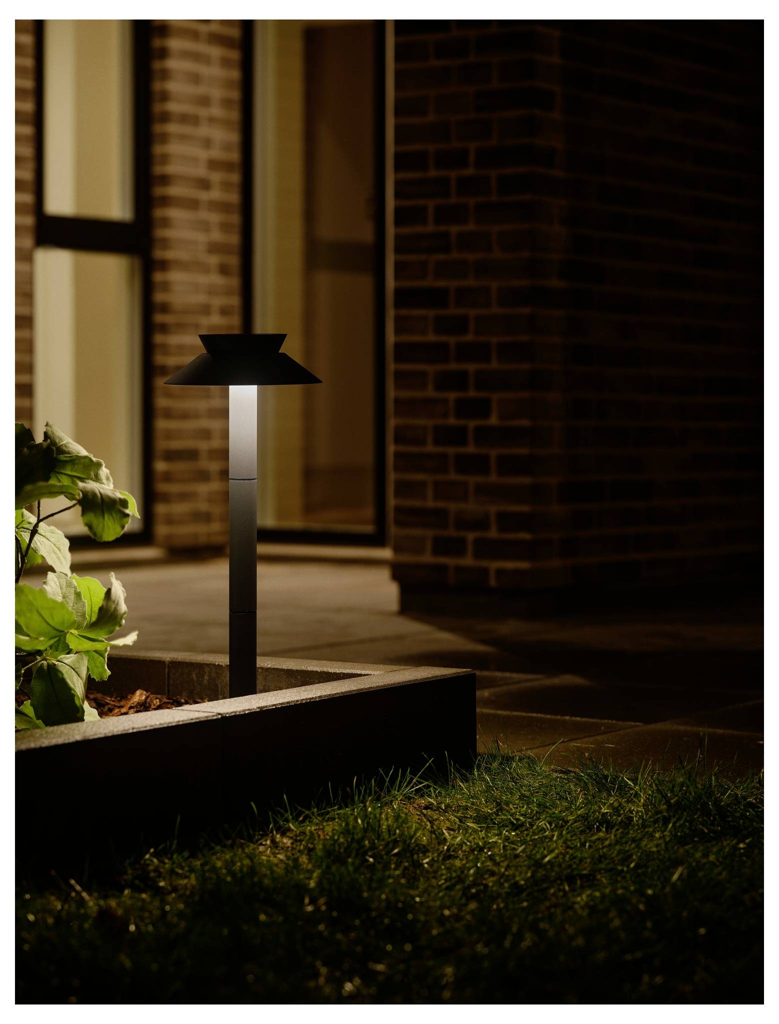 Ein moderner Außenlampenmast beleuchtet sanft einen Gartenweg neben einem Ziegelhaus in der Nacht und spendet subtile Beleuchtung für die Umgebung.
