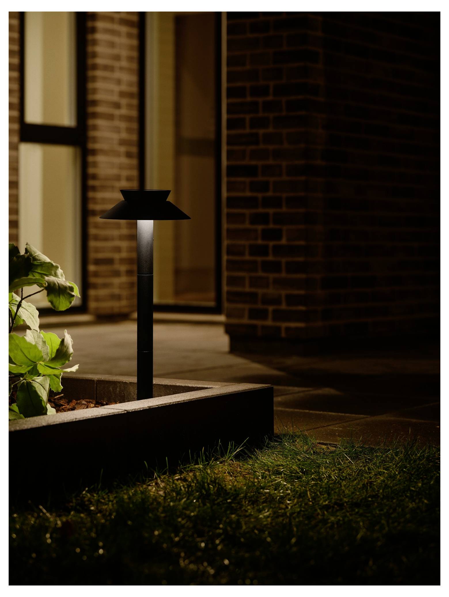 Eine kleine Gartenlampe beleuchtet sanft einen Weg neben einem Gebäude mit Backsteinmauern in der Nacht und hebt die nahegelegenen Pflanzen und das Gras hervor.