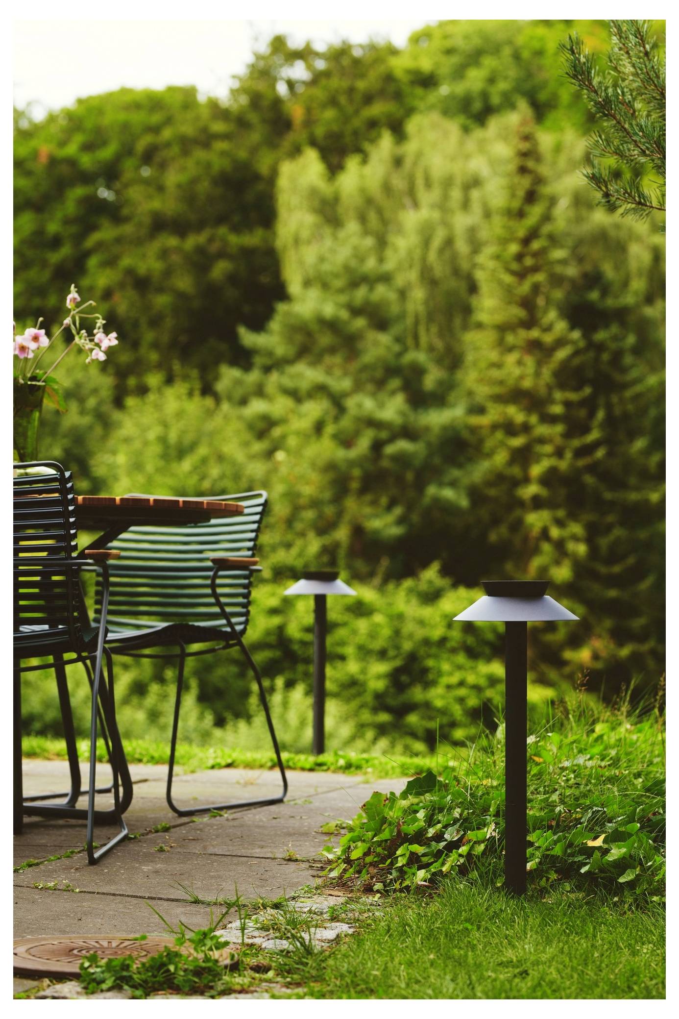 Eine Terrassenszene mit einem Tisch, Stühlen und Blumen mit Blick auf einen üppigen Garten mit verschiedenen Arten von Grünflächen und kleinen Laternen.