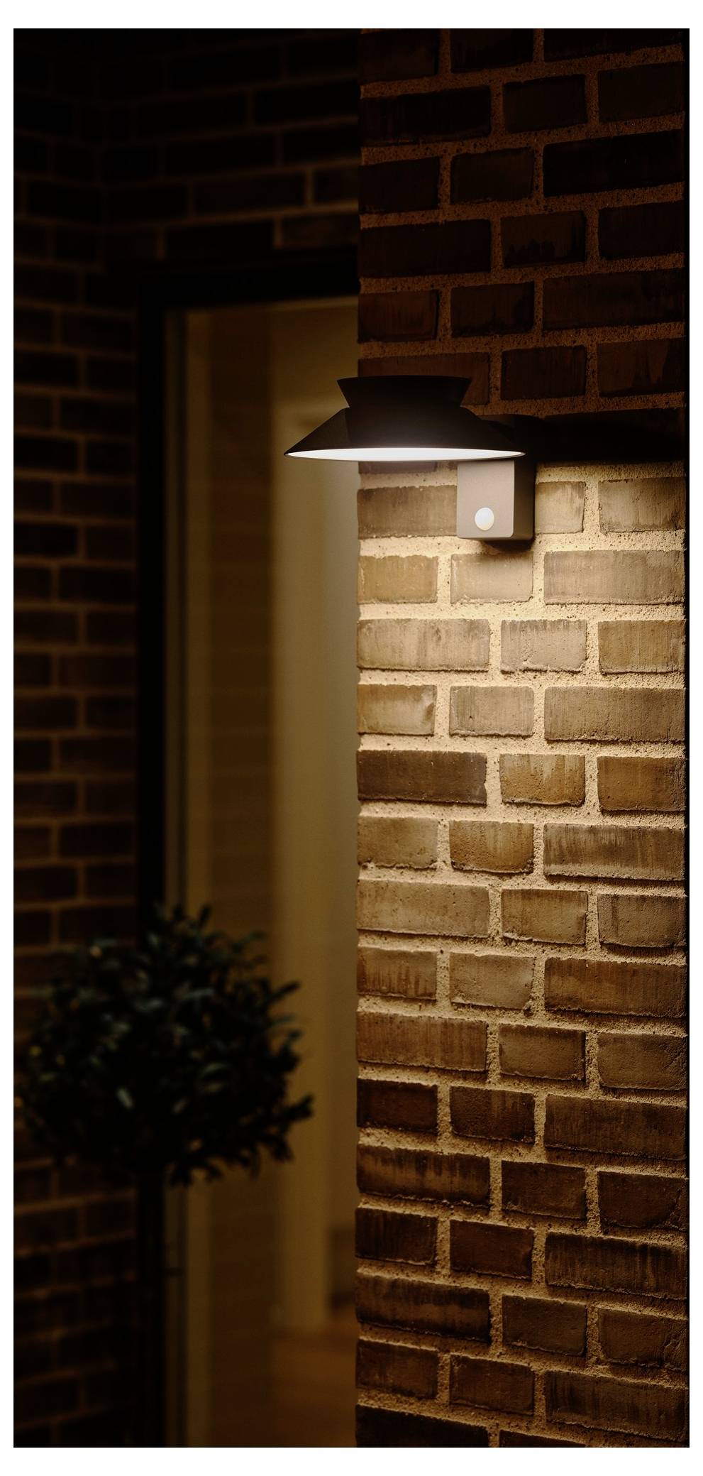 Eine Außenwandleuchte beleuchtet eine Backsteinwand neben einer offenen Tür und schafft eine warme, einladende Atmosphäre. Eine kleine Pflanze ist neben der Tür sichtbar.