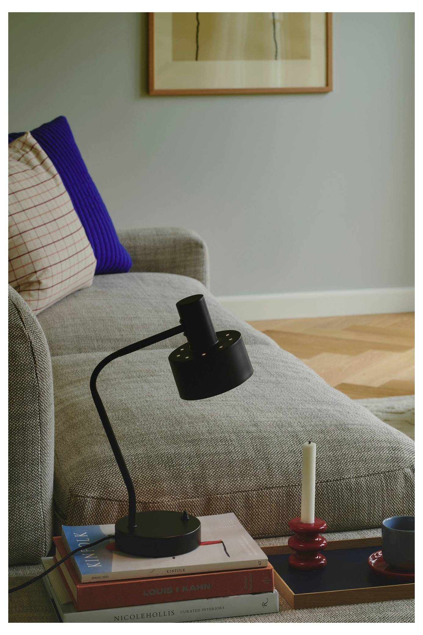 Ein gemütliches Wohnzimmerszenario mit einem grauen Sofa, das mit einem blauen Kissen verziert ist. Eine schwarze Schreibtischlampe steht auf einem Stapel von Büchern auf dem Beistelltisch.