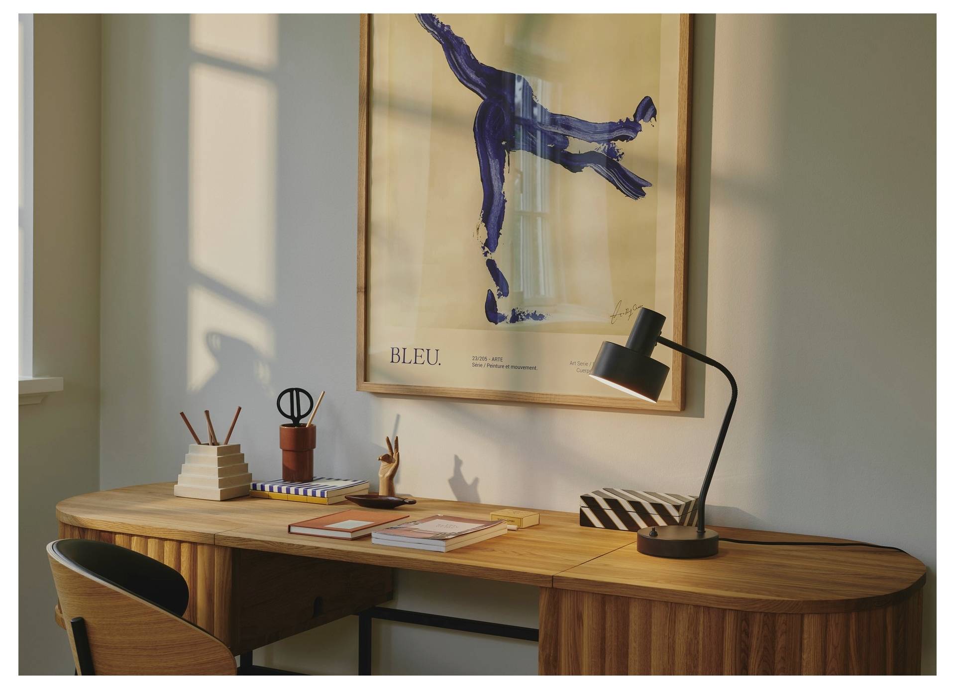 Ein Holzschreibtisch mit Büchern, Notizblöcken und einer Lampe in einem warm beleuchteten Raum. Ein Poster mit dem Titel „BLEU