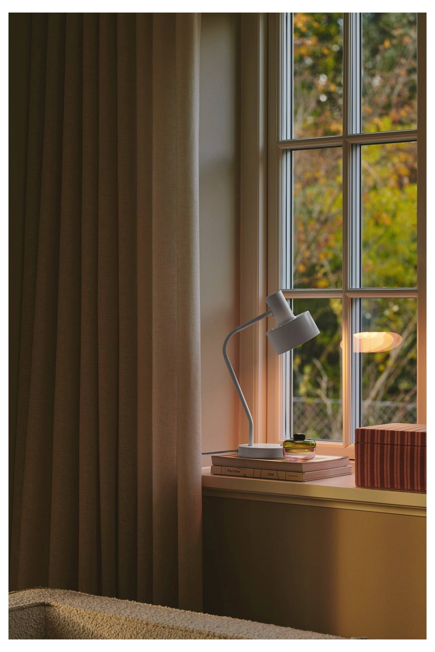 Eine gemütliche Ecke eines Zimmers mit einer Lampe auf der Fensterbank, Büchern und einem Blick auf Bäume durch das Fenster, das eine friedliche Leseecke andeutet.