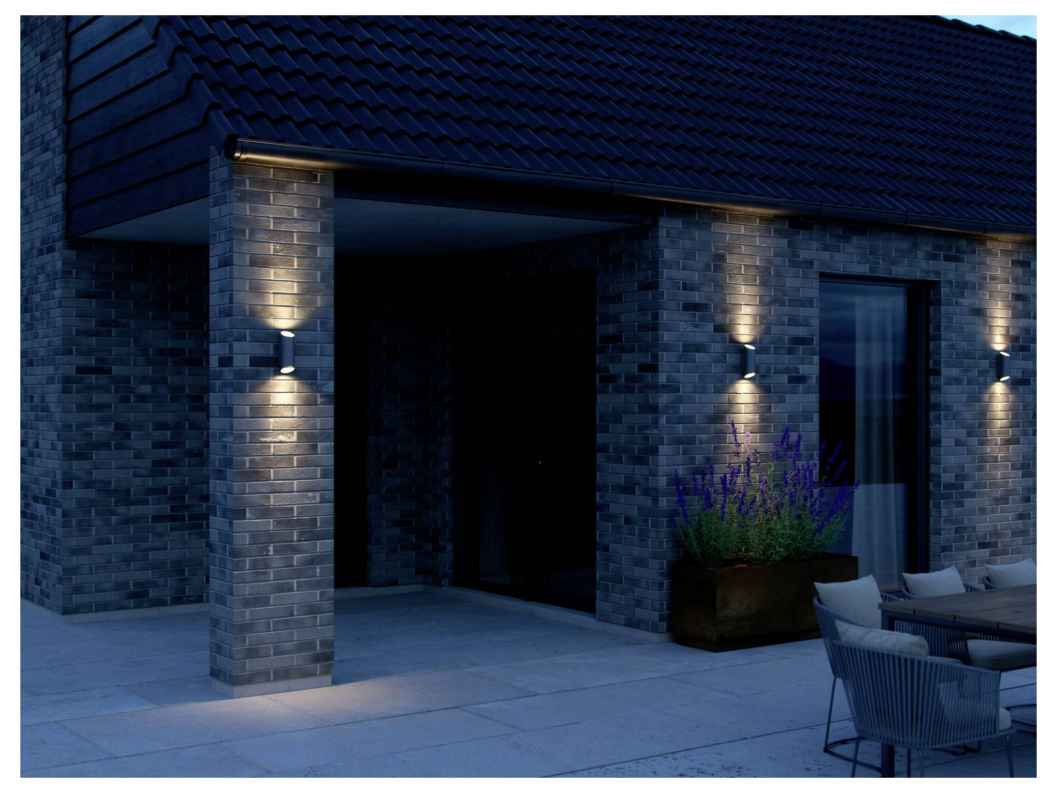 Modernes Hausäußeres in der Dämmerung mit zwei Wandleuchten, die eine Backsteinfront beleuchten. Ein Pflanzgefäß mit Lavendel steht in der Nähe auf der Terrasse.