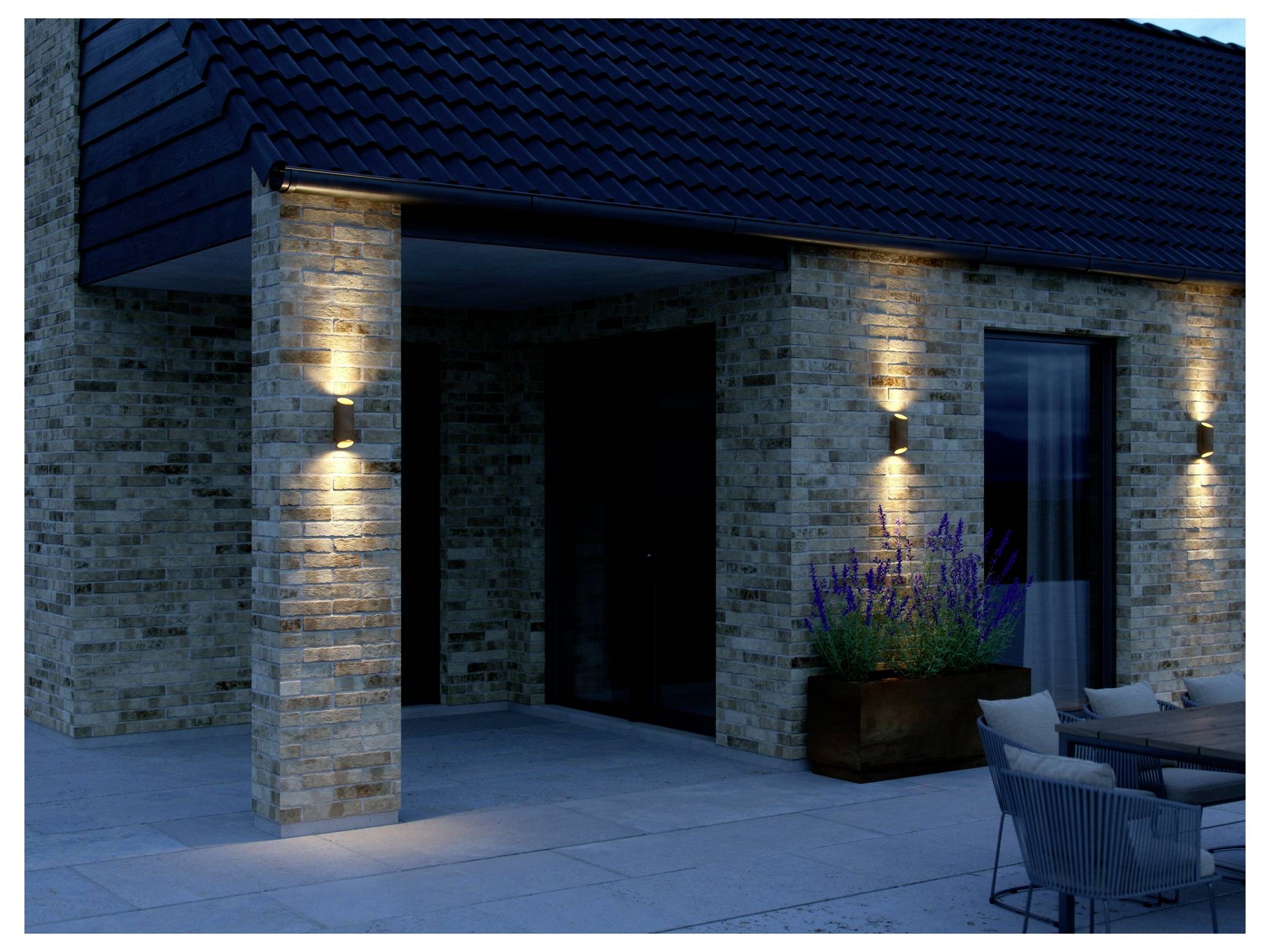 Ein modernes Hausäußeres mit Ziegelmauern und beleuchteten Wandleuchten am Eingang. Ein Blumenkübel mit violetten Blüten und Terrassenmöbel befinden sich in der Nähe.