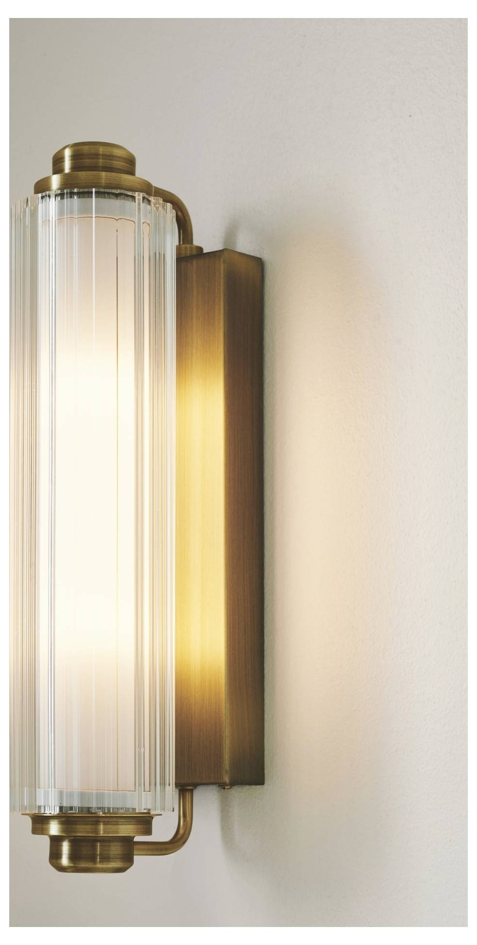 Eine zylindrische Wandleuchte mit geripptem Glas und Messingapplikationen gibt warmes, weiches Licht ab und ist an einer neutral gefärbten Wand montiert.