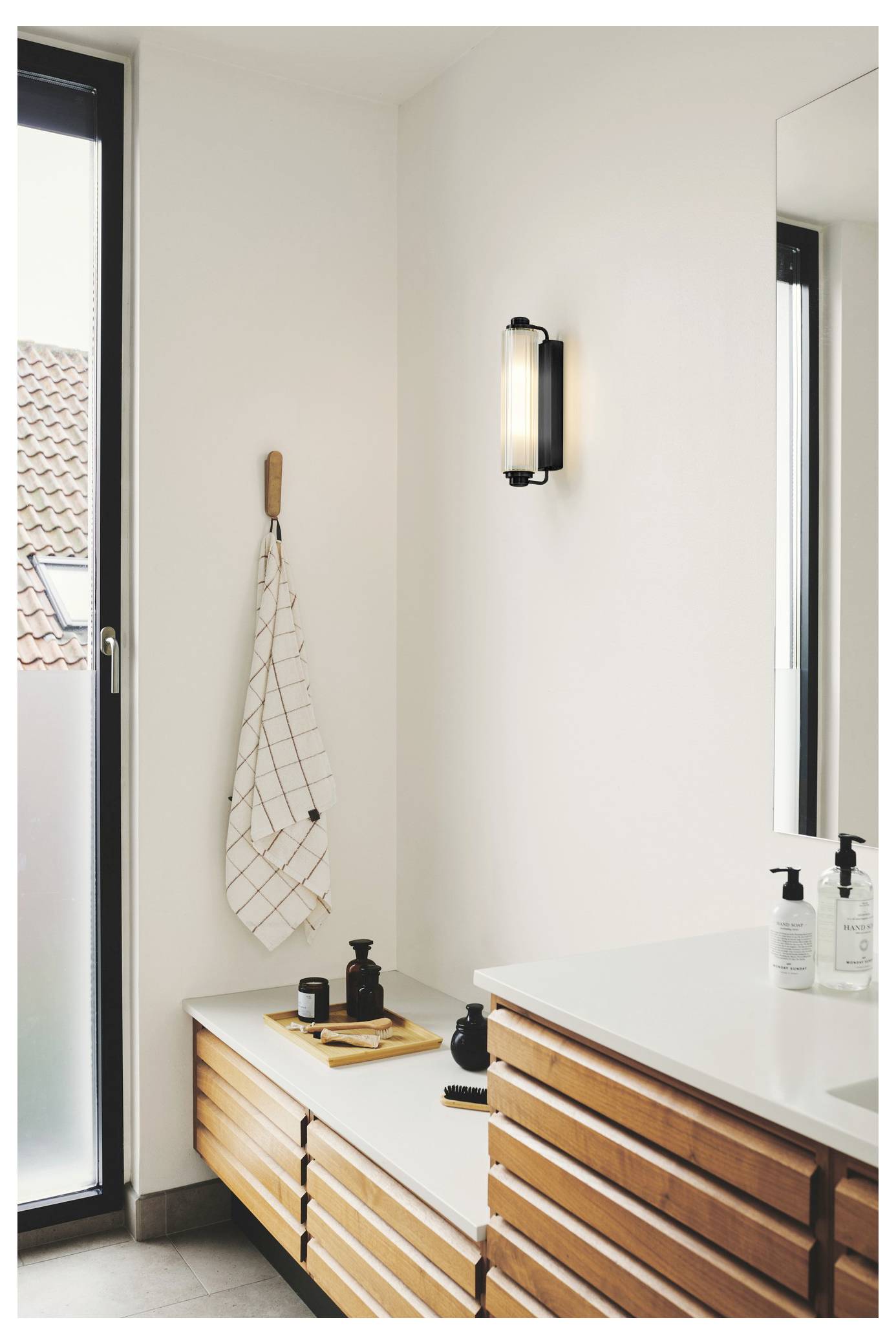 Modernes Badezimmer mit Holzschränken, weißer Arbeitsplatte, an der Wand montierter Leuchte oben, hängendem kariertem Handtuch und Glastür nach außen.