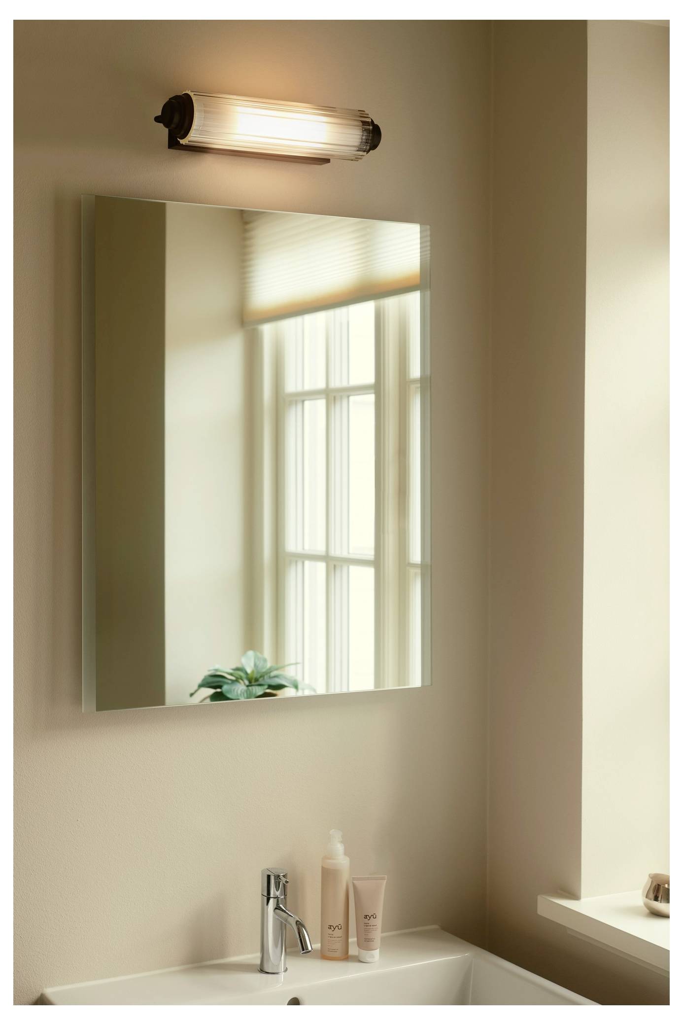 Ein Badezimmerschrank mit einem rechteckigen Spiegel, einer Wandleuchte darüber und einem Waschbecken darunter. Zwei Hautpflegeflaschen und eine kleine Pflanze stehen auf der Arbeitsfläche.