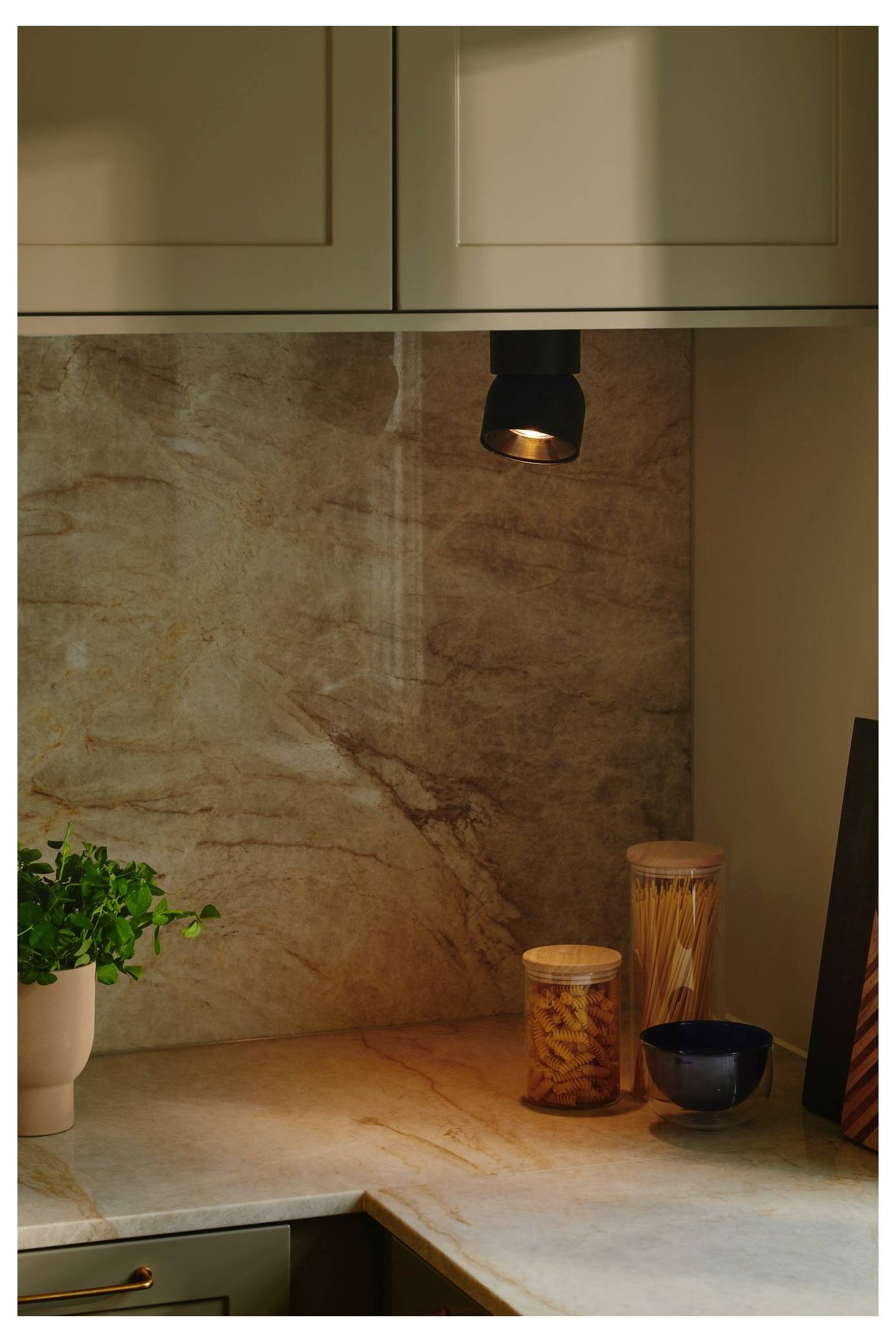 Eine gemütliche Küchenecke mit einem Scheinwerfer, der ein Glas Nudeln, eine kleine Pflanze und eine Schüssel auf einer Marmorarbeitsplatte unter Schränken beleuchtet.