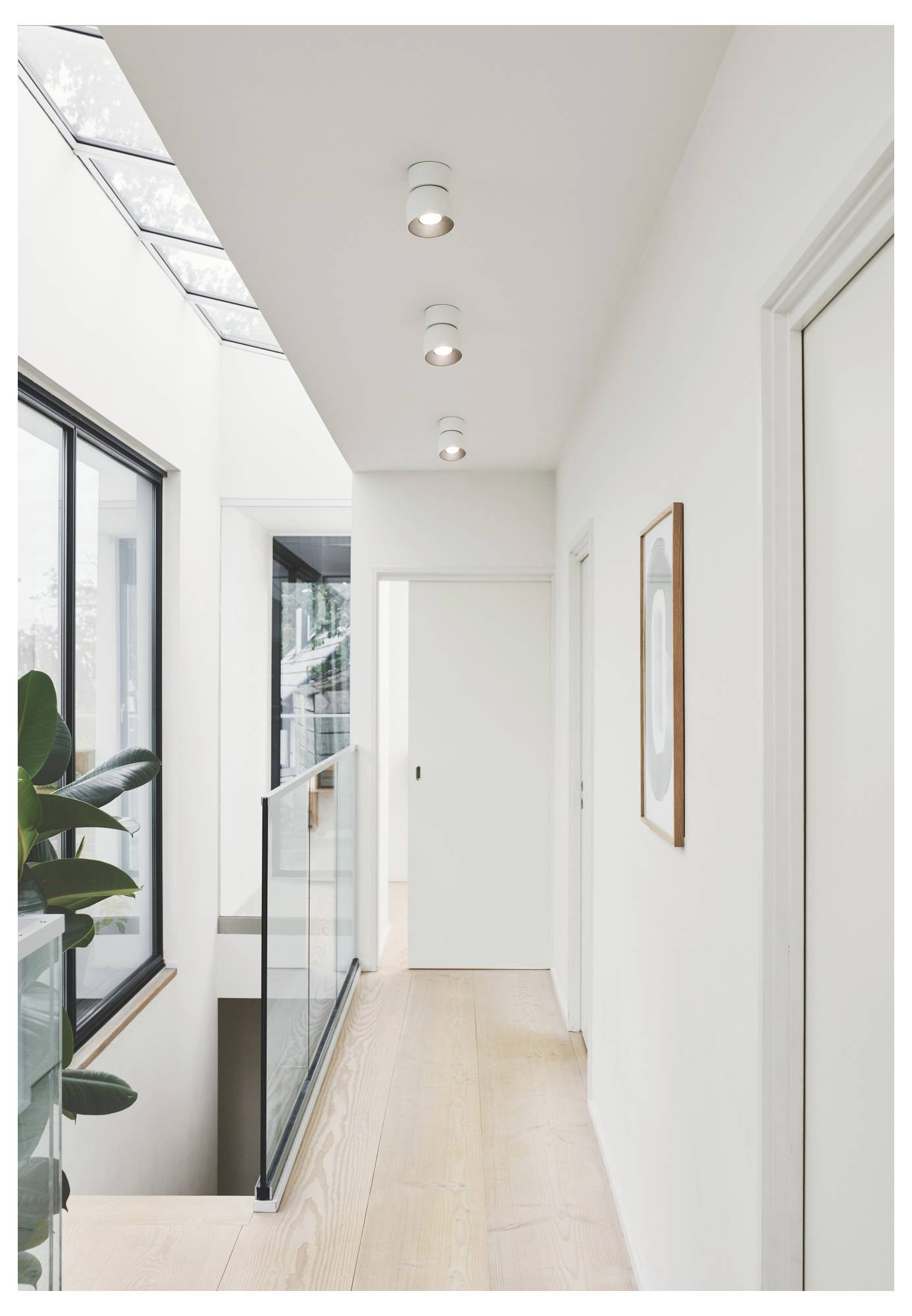 Ein moderner, heller Flur mit Holzfußboden, Glasgeländer, weißen Wänden, Deckenleuchten und einem großen Fenster auf der linken Seite.