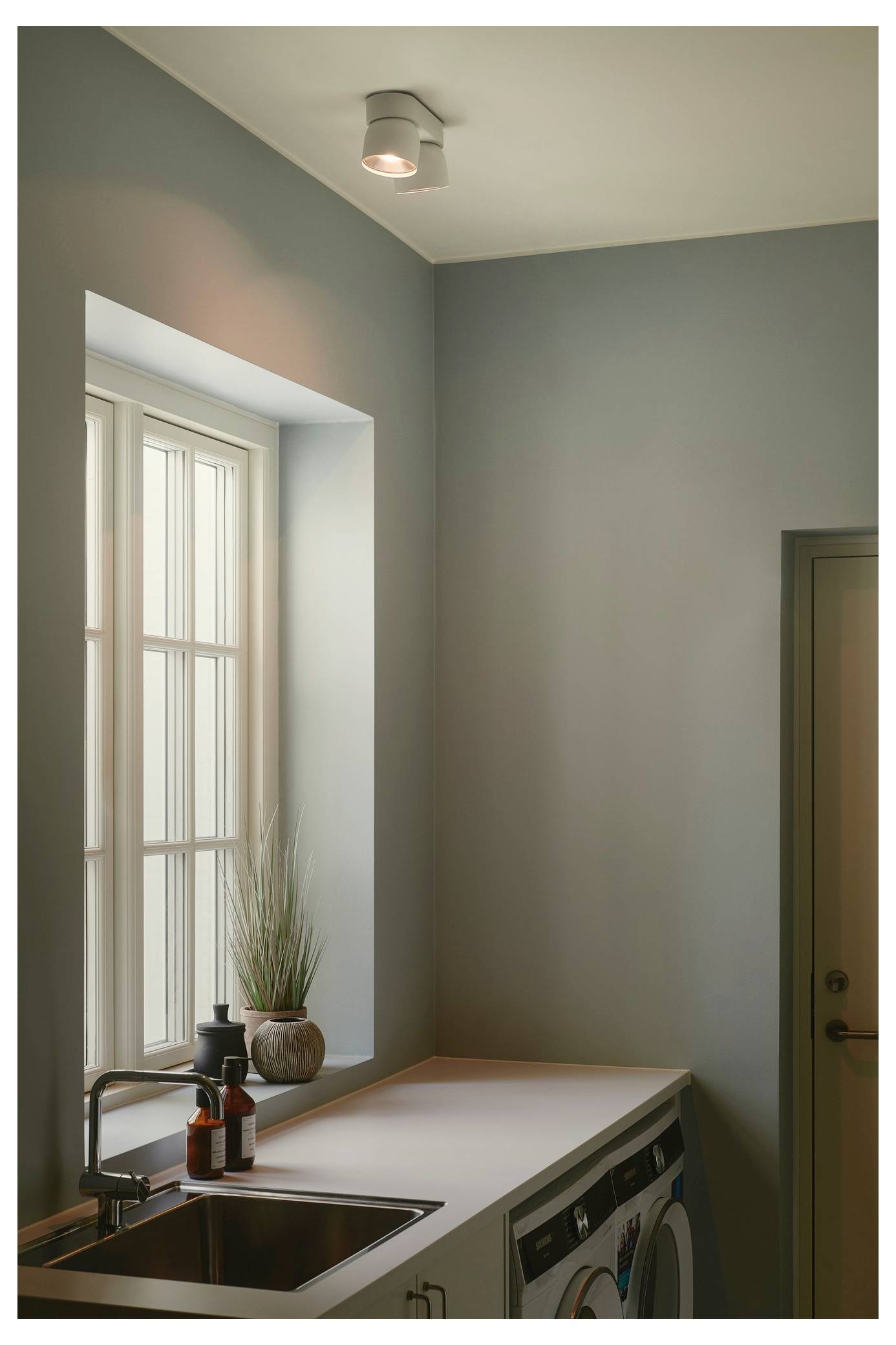 Waschküche mit einem Fenster, Waschmaschine und Trockner. Getopfte Pflanze und Seifenflaschen auf der Theke neben dem Waschbecken.