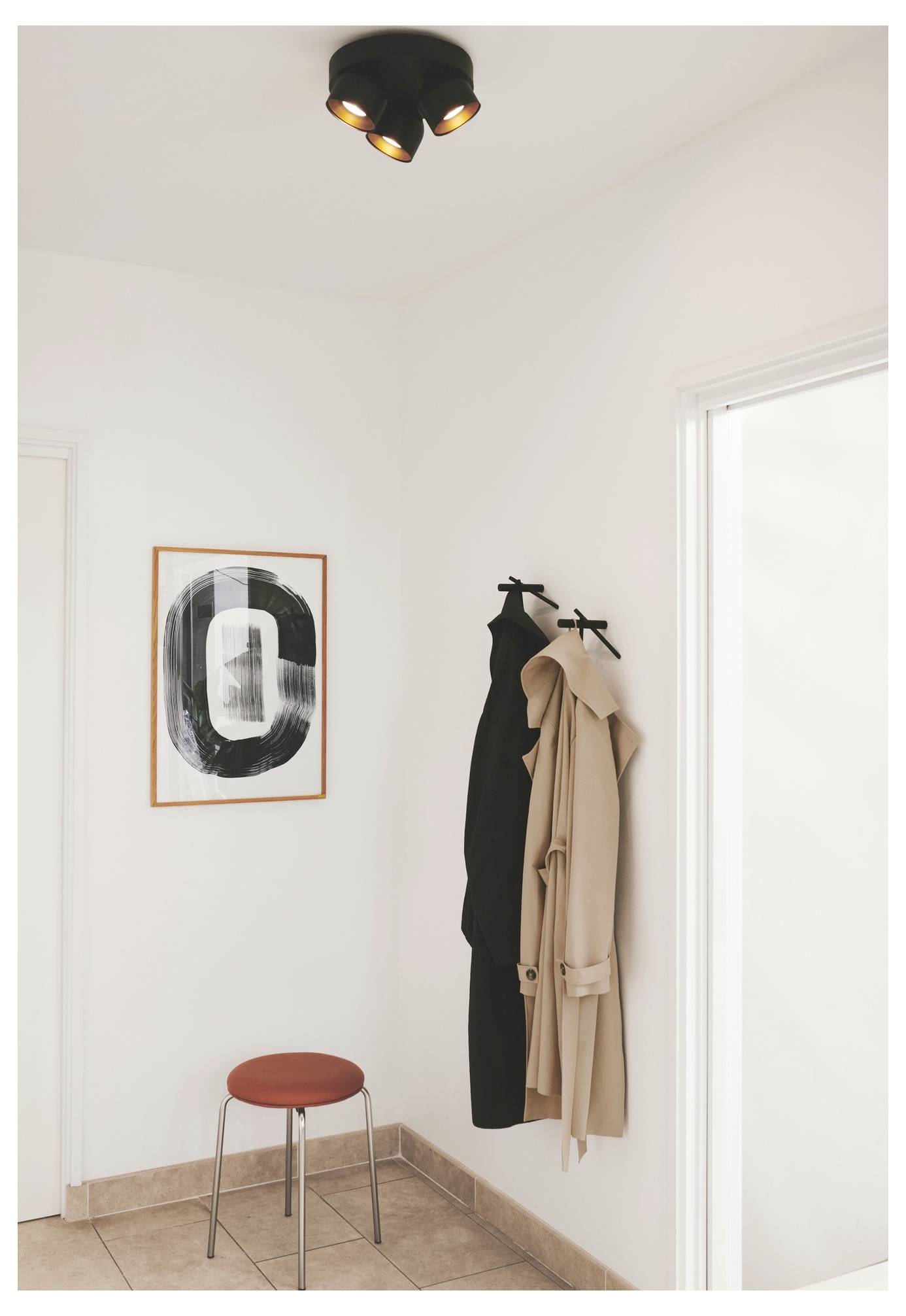 Eine minimalistische Flureck mit einem Hocker, zwei hängenden Mänteln und abstrakter Kunst an der Wand. Schwarze Deckenleuchte darüber.