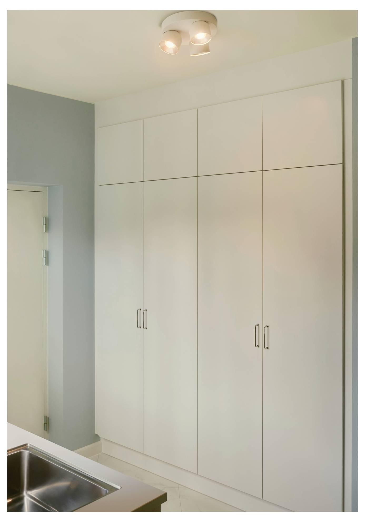 Hohe, weiße Küchenschränke mit eleganten Griffen, vor einer hellblauen Wand aufgestellt, neben einem Spülbecken und unter Deckenleuchten.