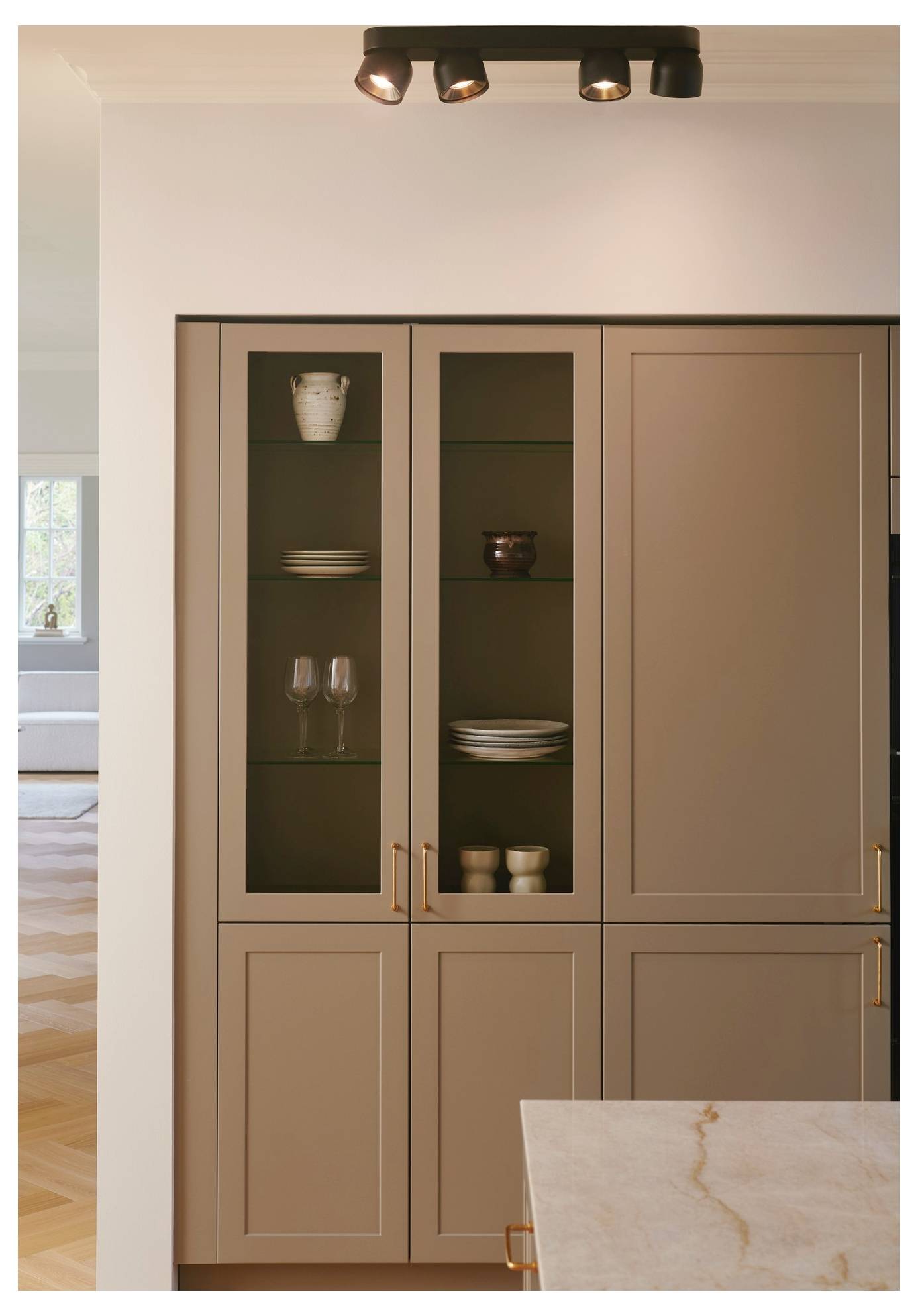 Eine moderne Küche mit beigen Schränken mit Glastüren, die Geschirr und Glaswaren ausstellen, unter einer Deckenleiste mit Strahler.