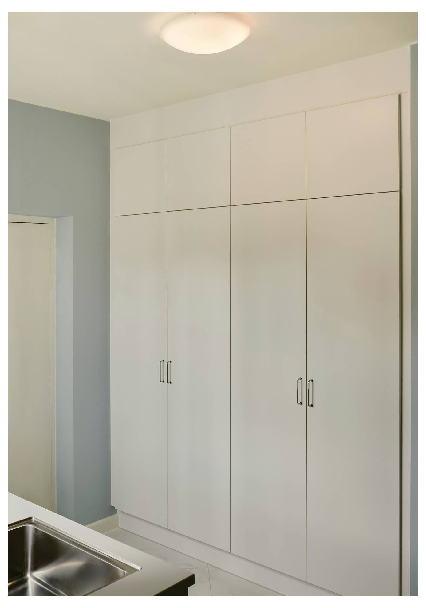 Hohe weiße Küchenschränke mit eleganten Metallgriffen, an einer hellblauen Wand positioniert, neben einem Edelstahlspülbecken.