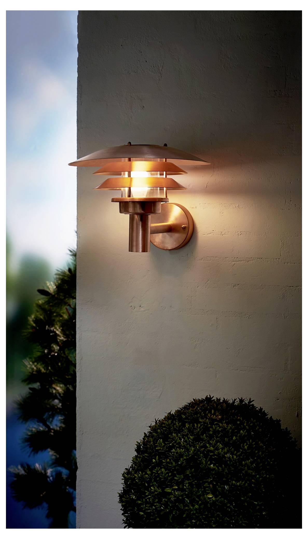 Eine Außenwandleuchte strahlt ein warmes Licht auf eine Betonmauer aus, mit unscharfem Grün im Hintergrund, das eine gemütliche Abendatmosphäre schafft.