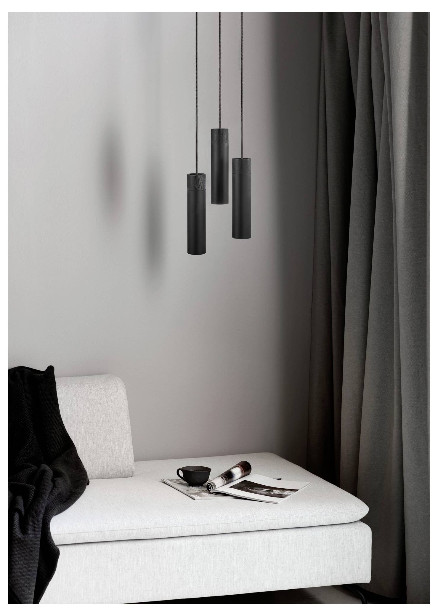 Eine moderne Wohnzimmerecke mit einer weißen Chaiselongue, schwarzen Pendelleuchten, geschlossenen Gardinen, einer schwarzen Decke und einer Zeitschrift mit einer Tasse.