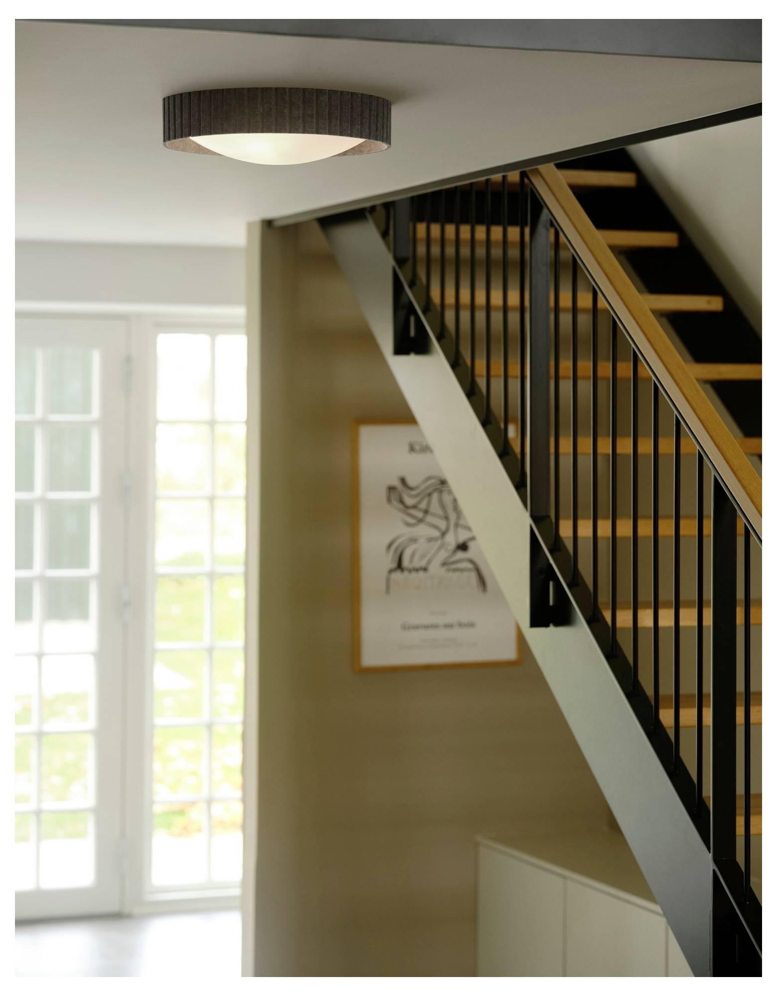 Ein modernes Interieur mit einer Holztreppe, schwarzem Geländer und einer runden Deckenleuchte. Ein Poster hängt an der Wand neben großen Fenstern.