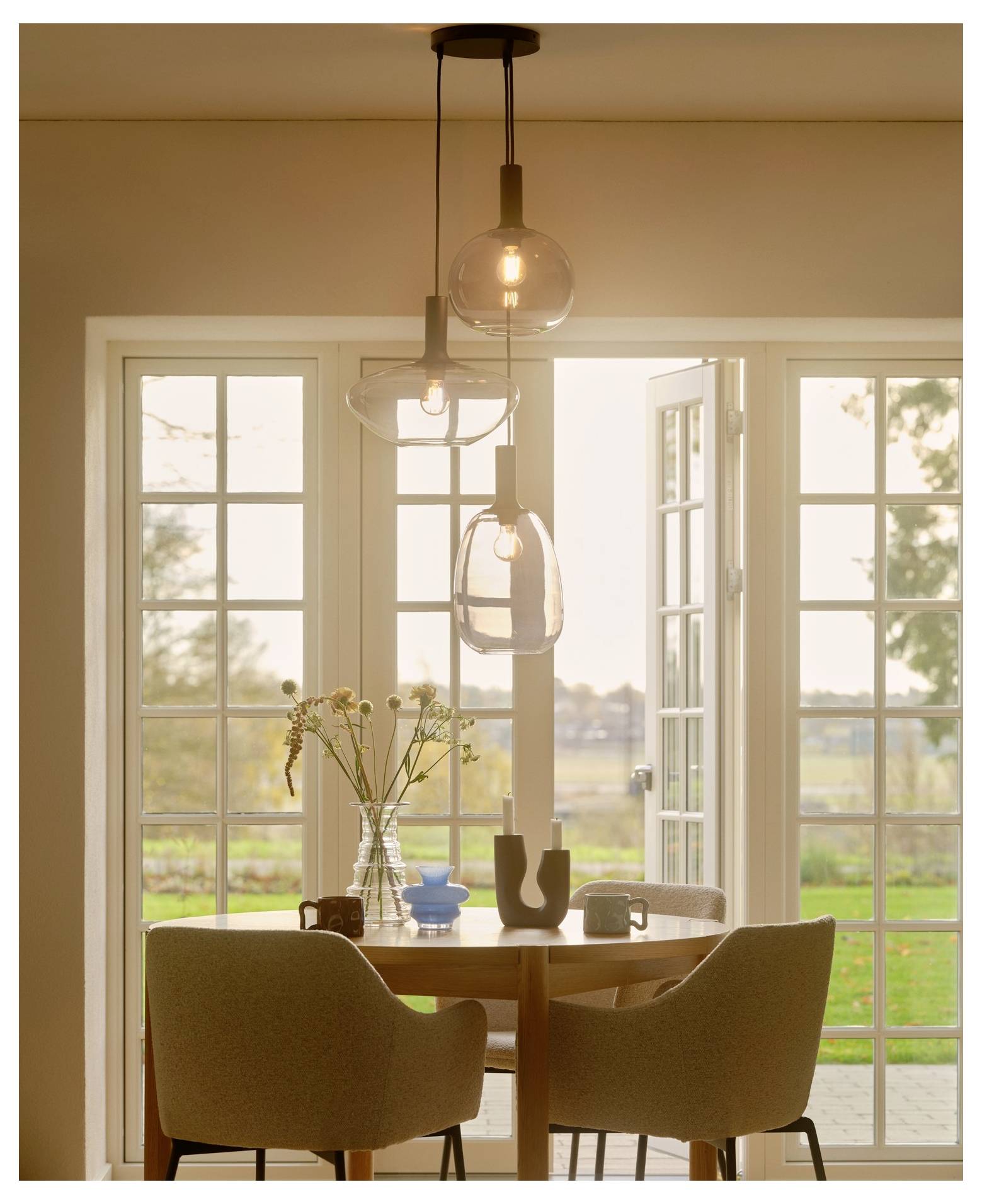 Ein gemütlicher Essbereich mit einem runden Tisch, zwei Stühlen und dekorativen Gegenständen. Große Fenster öffnen sich zu einer malerischen Aussicht auf einen Garten.