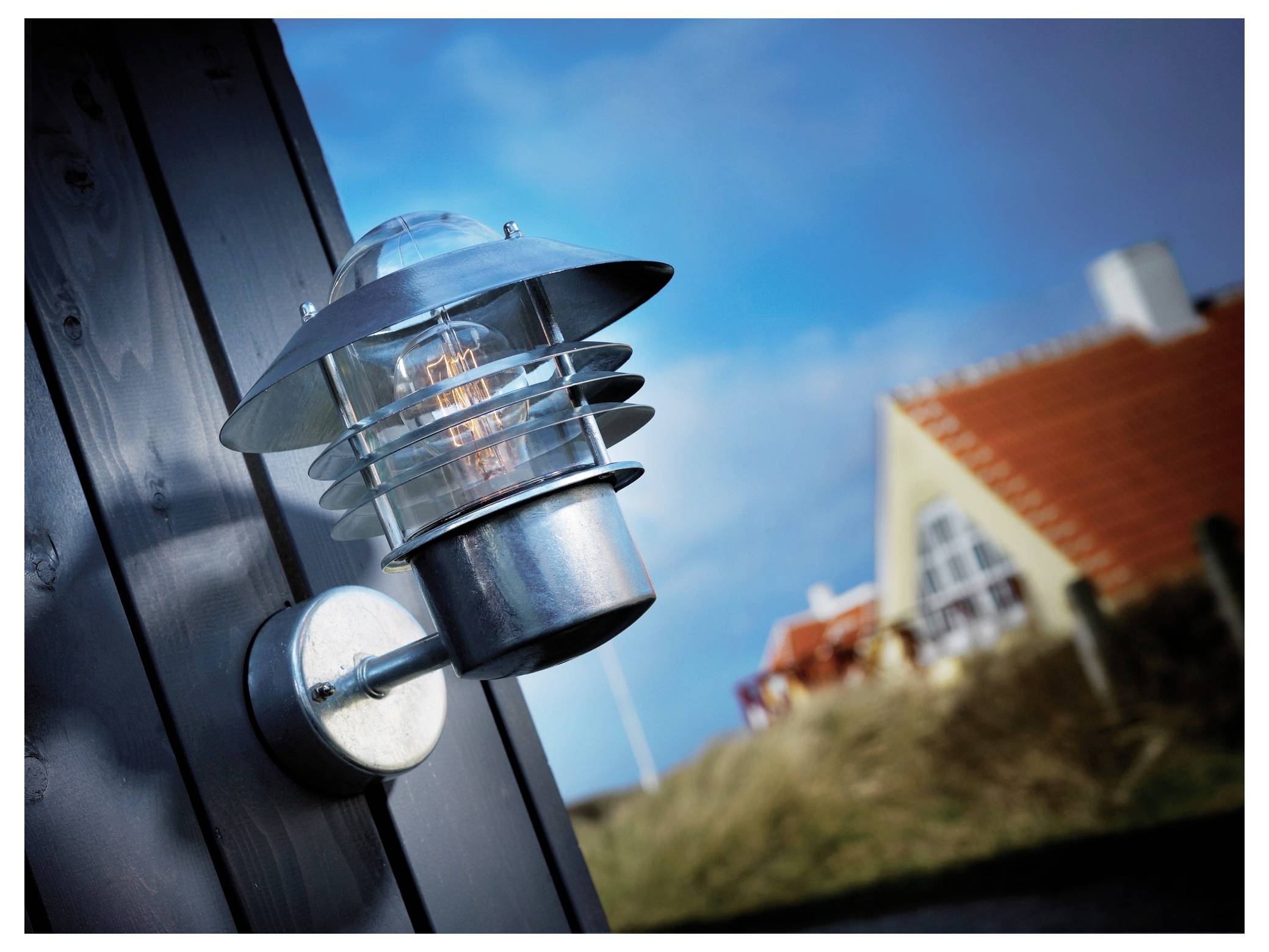Eine auf einer dunklen Wand montierte Außenleuchte, mit einem unscharfen Haus mit rotem Dach und blauem Himmel im Hintergrund.