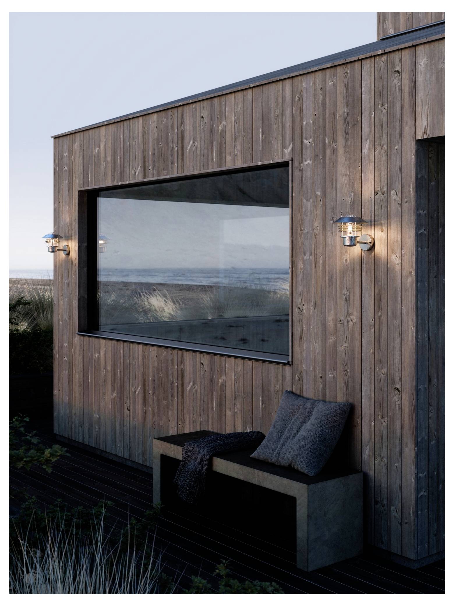 Eine moderne Holzhütte mit großen Glasfenstern mit Blick auf eine Küstenlandschaft, ausgestattet mit zwei Wandleuchten und einer Bank mit Kissen im Freien.