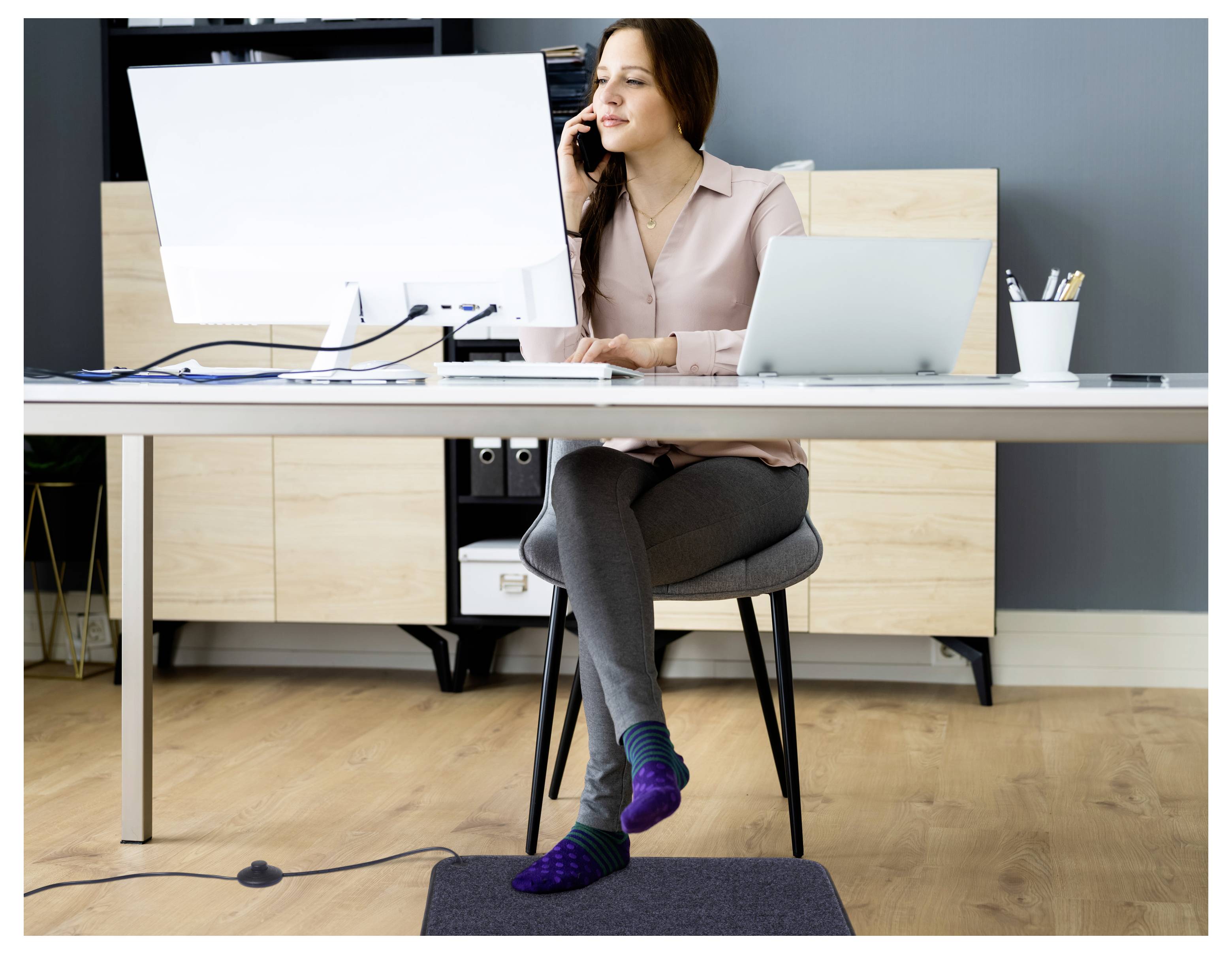Frau sitzt an einem Schreibtisch in einem Büro, telefoniert, während sie an einem Computer arbeitet, mit einem Laptop neben ihr.