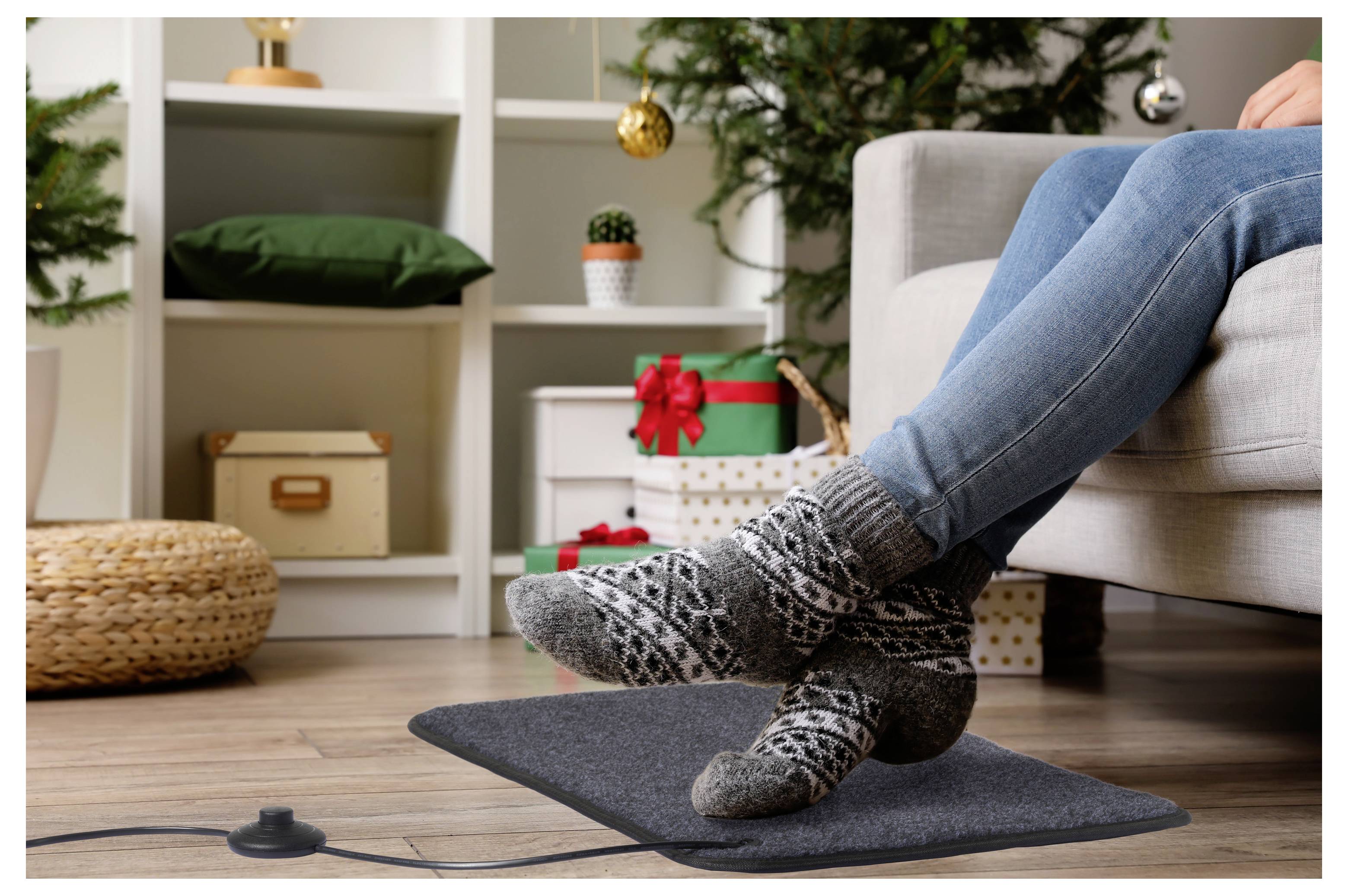 Person sitzt auf einem Sofa und trägt gemusterte Wollsocken, entspannt sich mit den Füßen auf einer beheizten Fußmatte in einem gemütlichen Wohnzimmer mit Feiertagsdekoration.