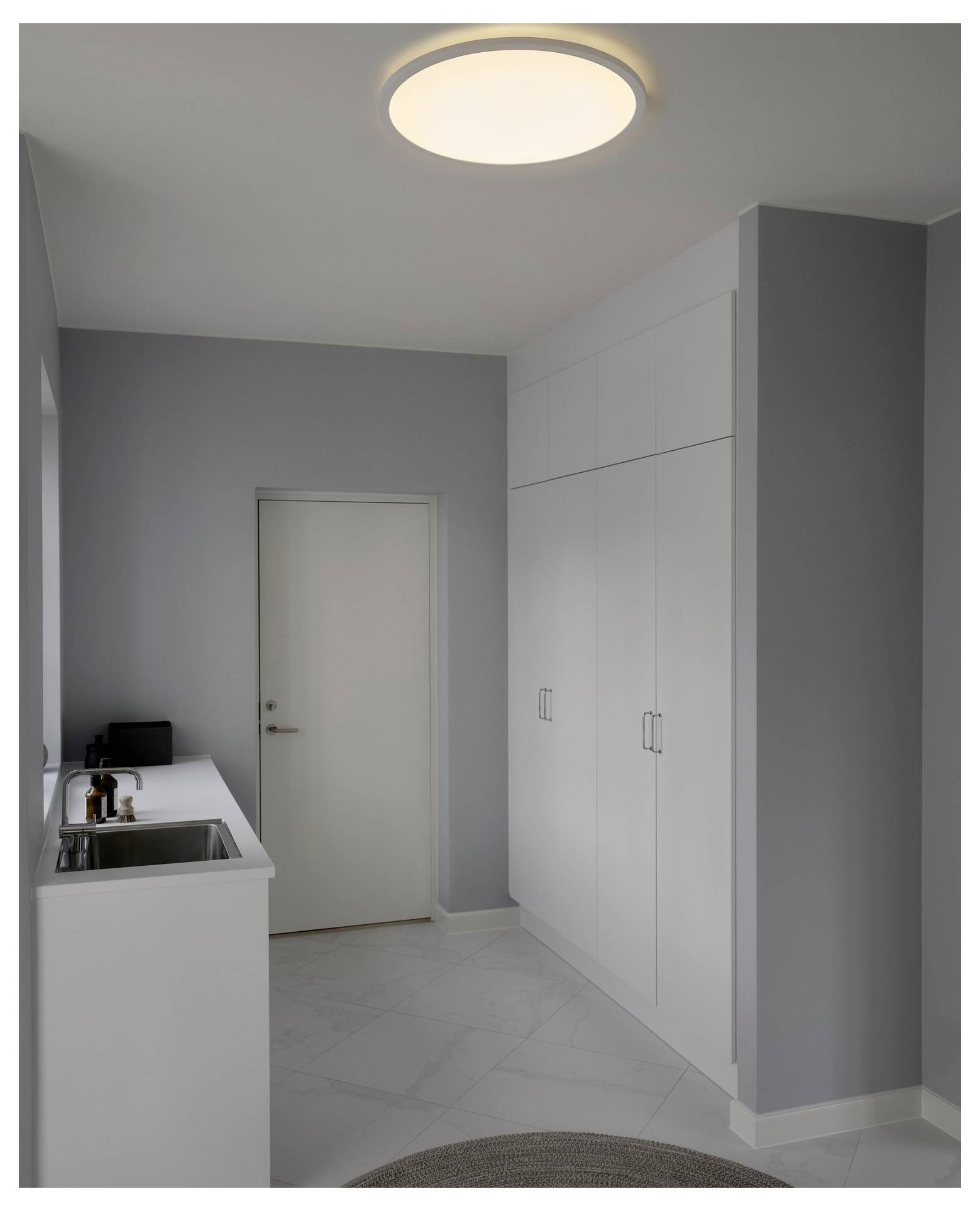 Ein minimalistisches Waschzimmer mit grauen Wänden, weißen Schränken und einer runden Deckenleuchte. Ein Waschbecken und eine Arbeitsplatte sind auf der linken Seite.