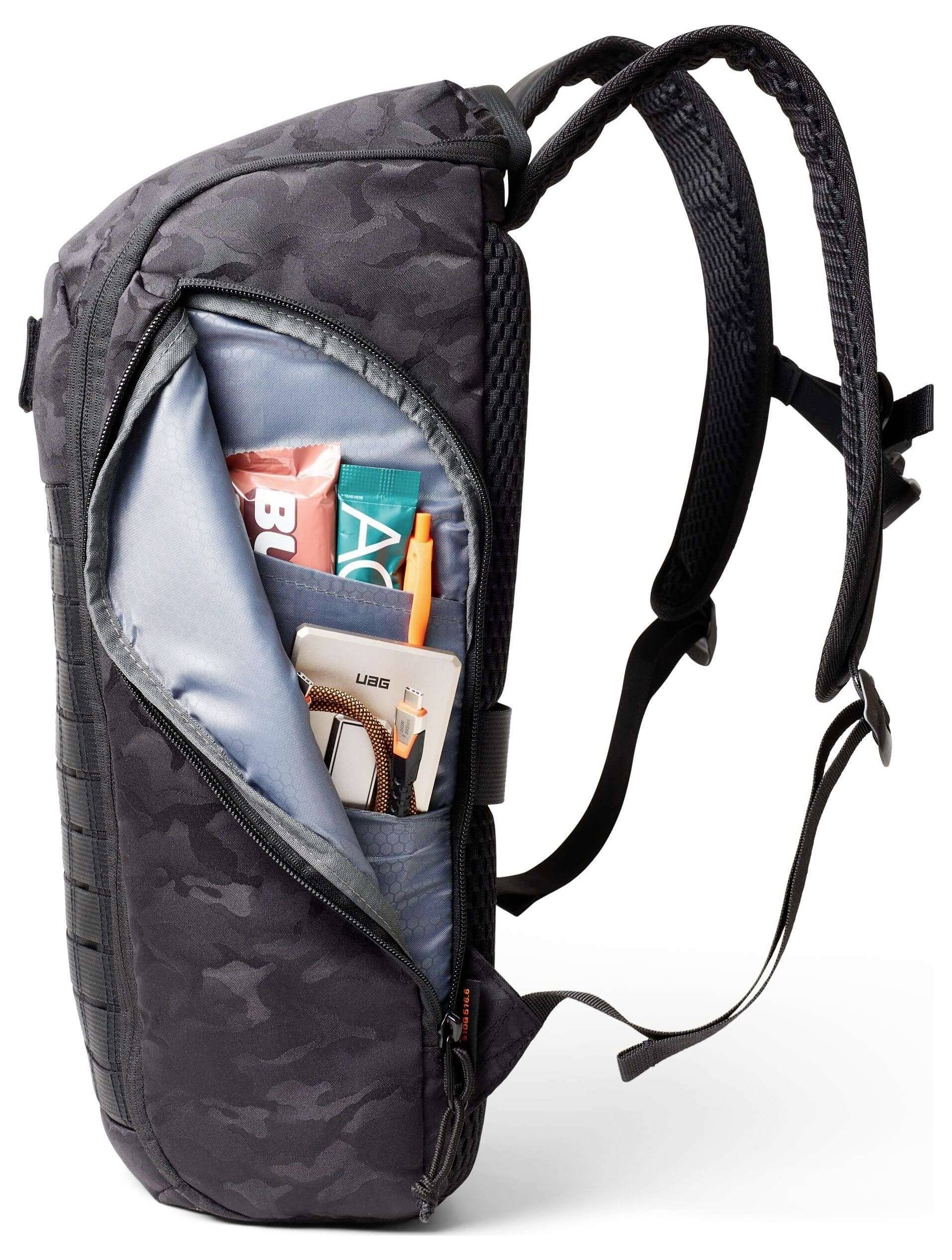 Eine Seitenansicht eines schwarzen Rucksacks mit Camouflage-Muster und einer offenen Tasche, aus der Magazine, ein Buch und ein Telefon herausschauen.