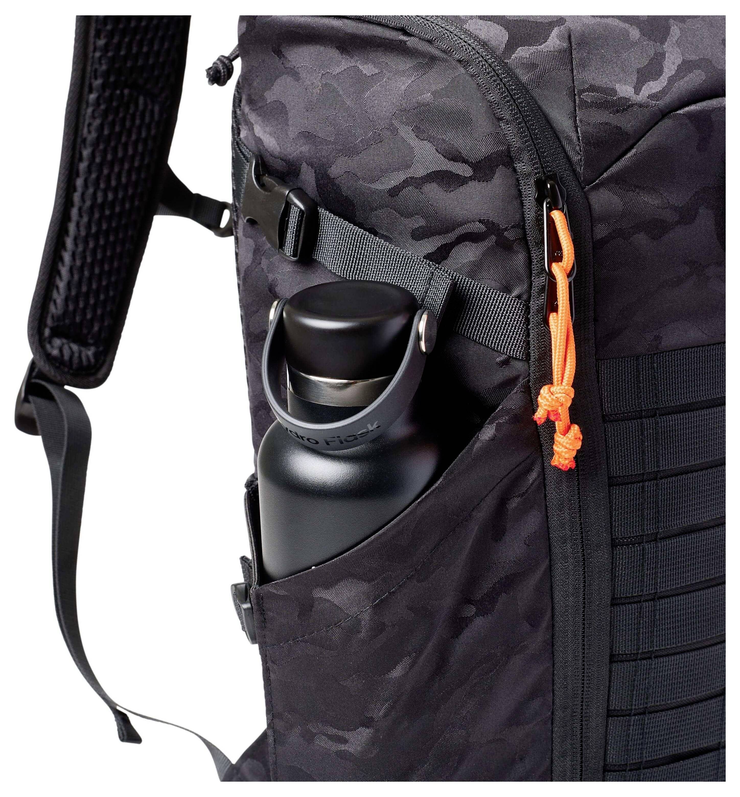 Ein schwarzer Tarnrucksack mit einem orangefarbenen Reißverschluss-Anhänger, der eine schwarze Wasserflasche in seiner Seitentasche hält, was seine Speicherkapazität hervorhebt.