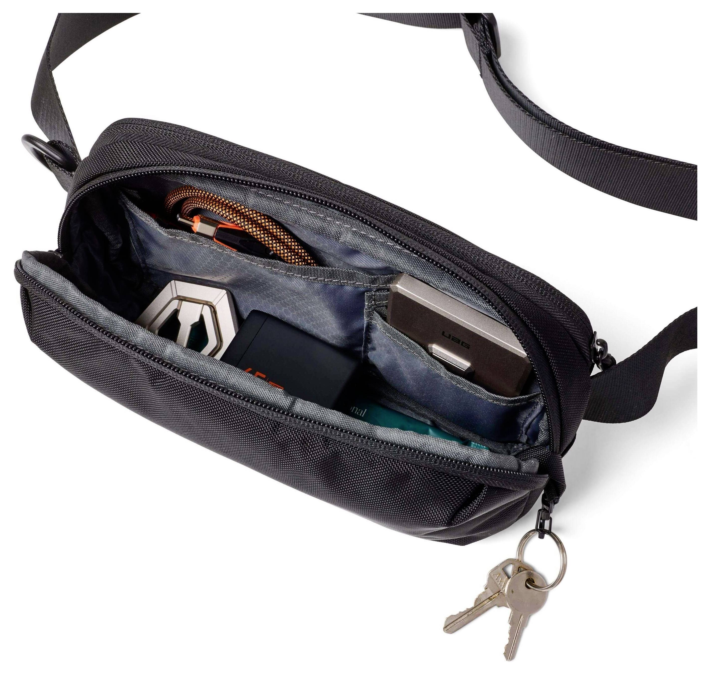 Eine schwarze Umhängetasche ist offen und zeigt ihren Inhalt, darunter einen kleinen Lautsprecher, ein tragbares Ladegerät und Schlüssel, die am Reißverschlussschieber befestigt sind.