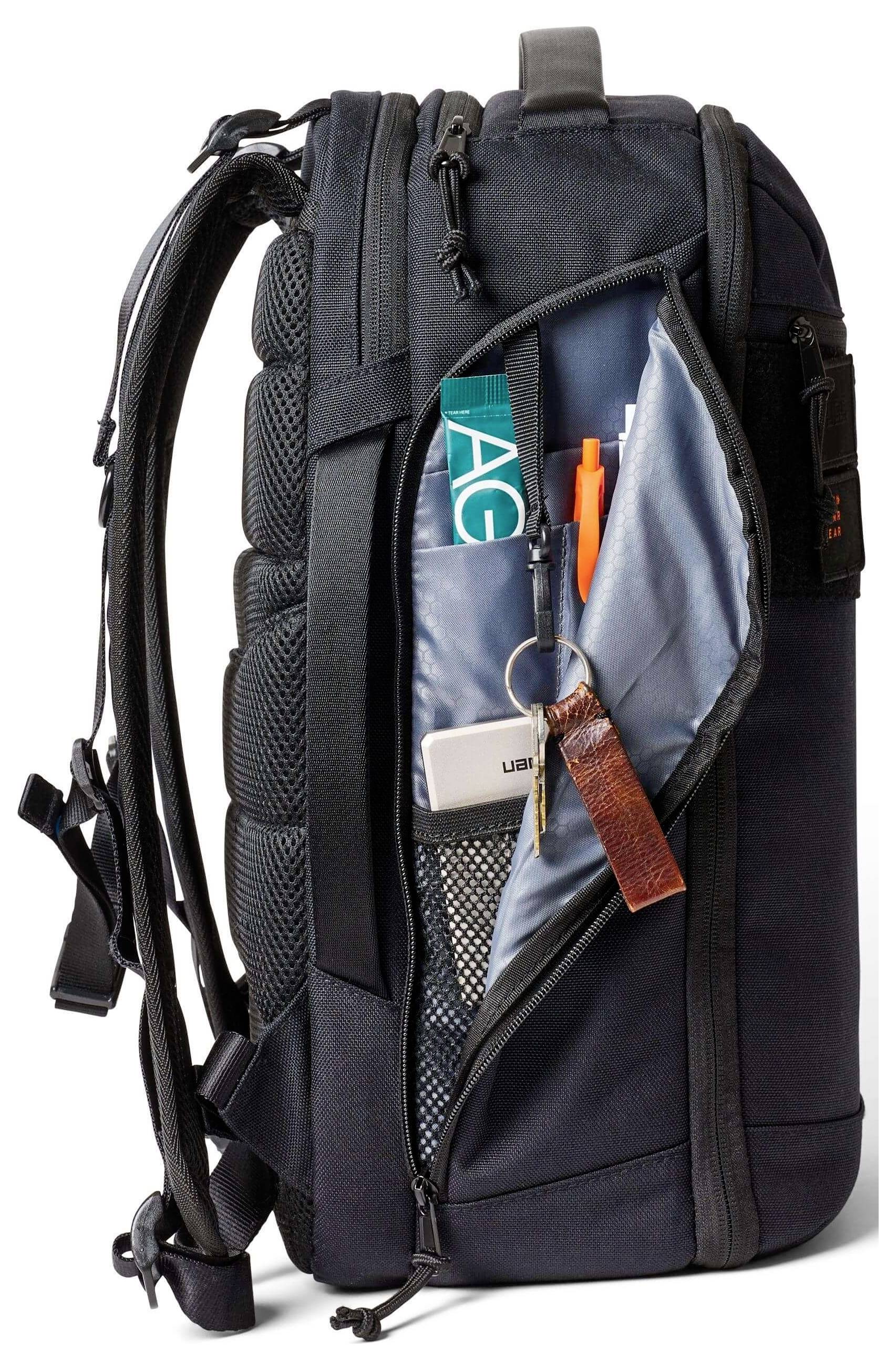 Ein schwarzer Rucksack, der vorne offen ist und organisierte Fächer zeigt, in denen sich ein Schlüsselanhänger, ein Stift, eine Zeitschrift und ein kleines Gerät befinden.