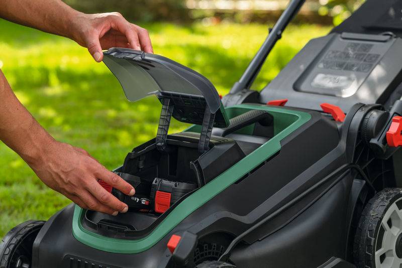 Eine Person setzt eine Batterie in einen schnurlosen Rasenmäher ein, der auf Gras in einer Gartenumgebung zu sehen ist. Das Batteriefach des Mähers ist offen.