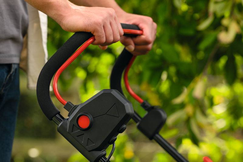 Eine Person hält den Griff eines elektrischen Rasenmähers, umgeben von Grünflächen, was auf eine Gartenarbeit im Freien hindeutet.