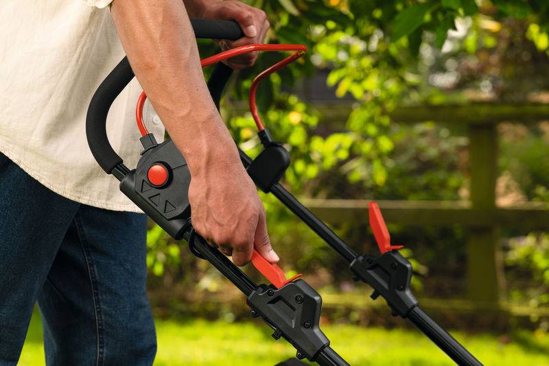 Eine Person stellt den Griff eines Rasenmähers in einer Gartenumgebung ein, mit üppigem grünem Laub und einem Holzzaun im Hintergrund.